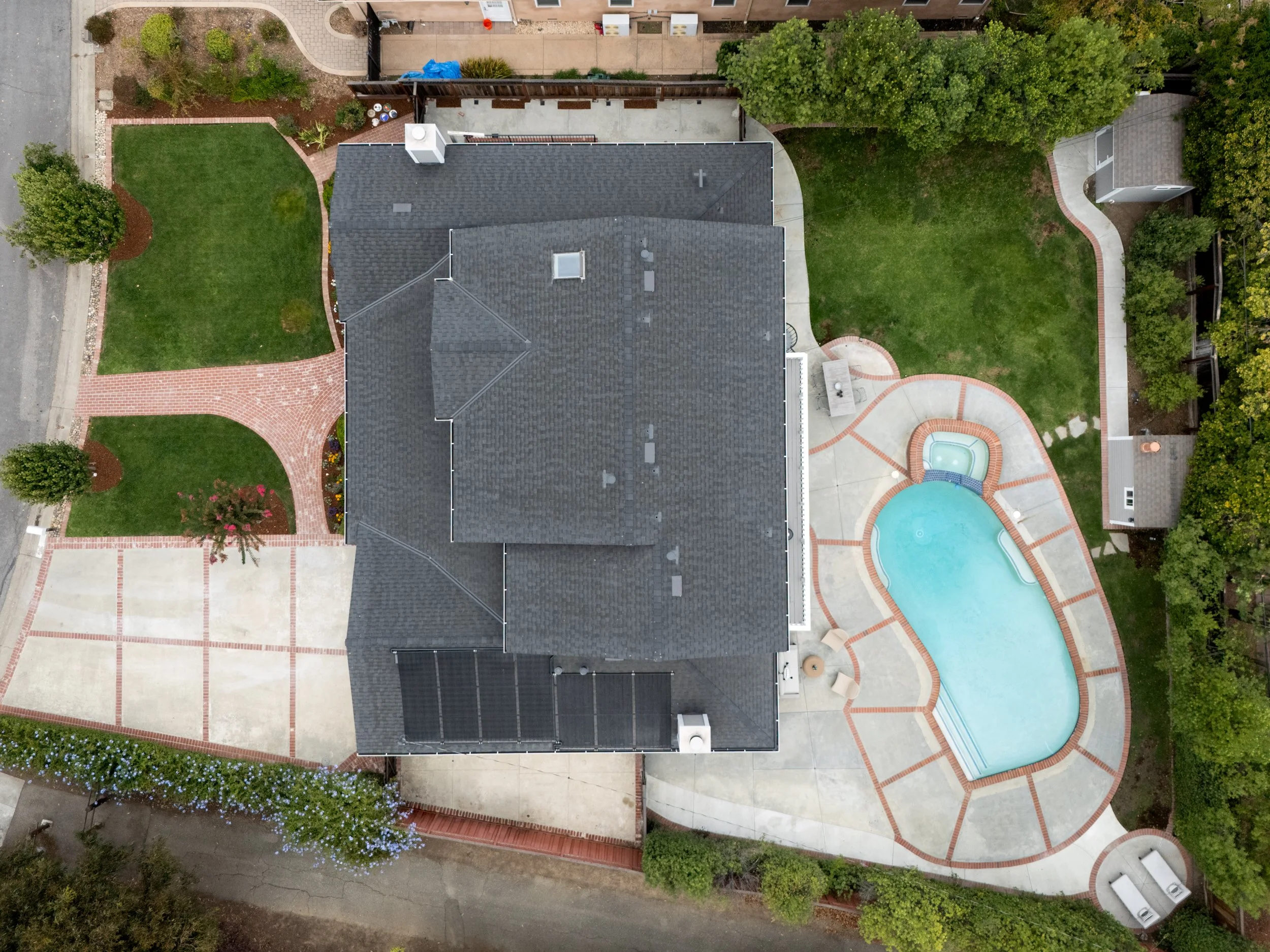 An aerial view of a backyard showing a house with a dark gray shingle roof, a swimming pool, a green lawn, brick pathways, and surrounding trees and plants.