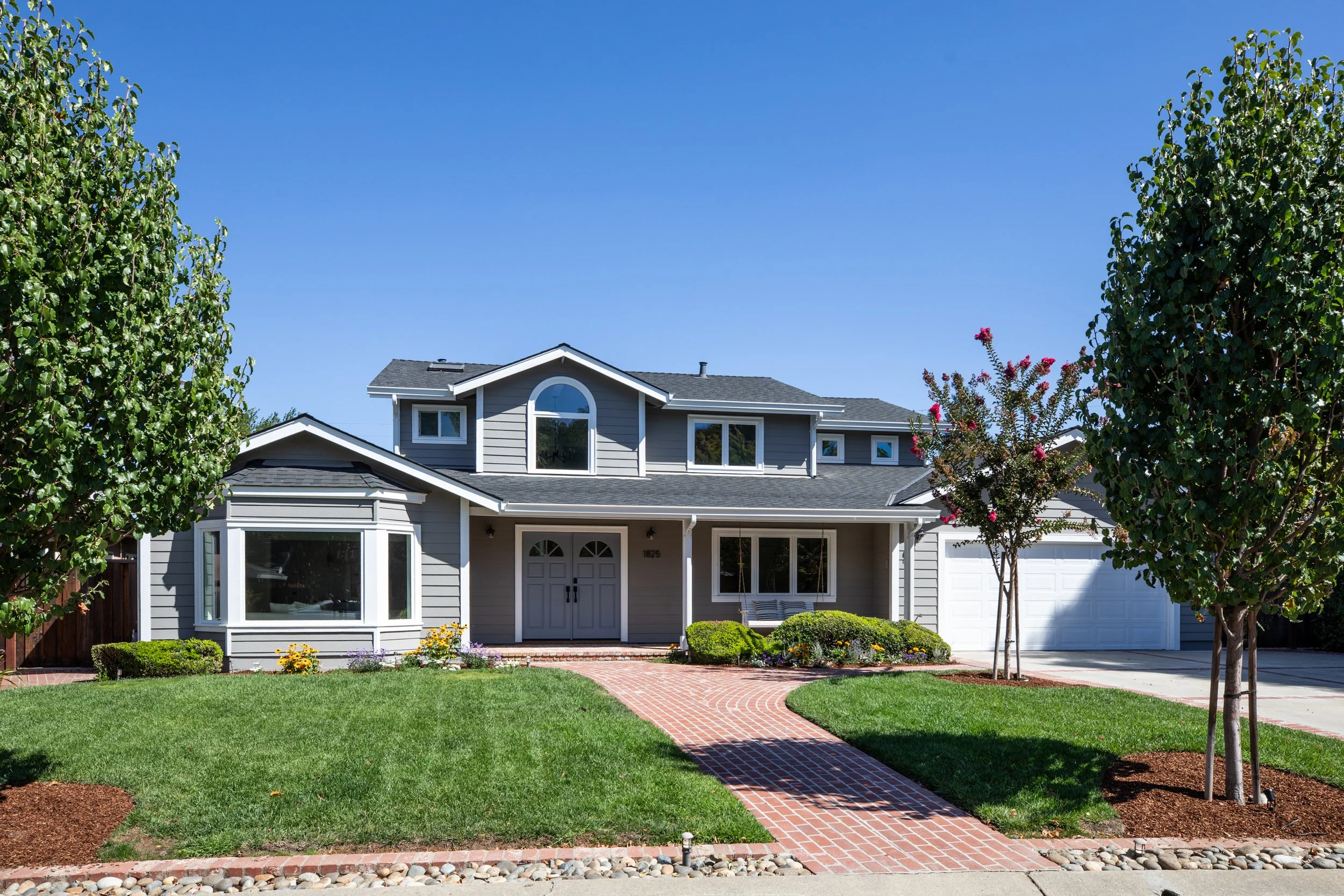 Front view of a modern two-story house with a well-maintained lawn, brick walkway, trees, and shrubs in a suburban neighborhood under a clear blue sky.