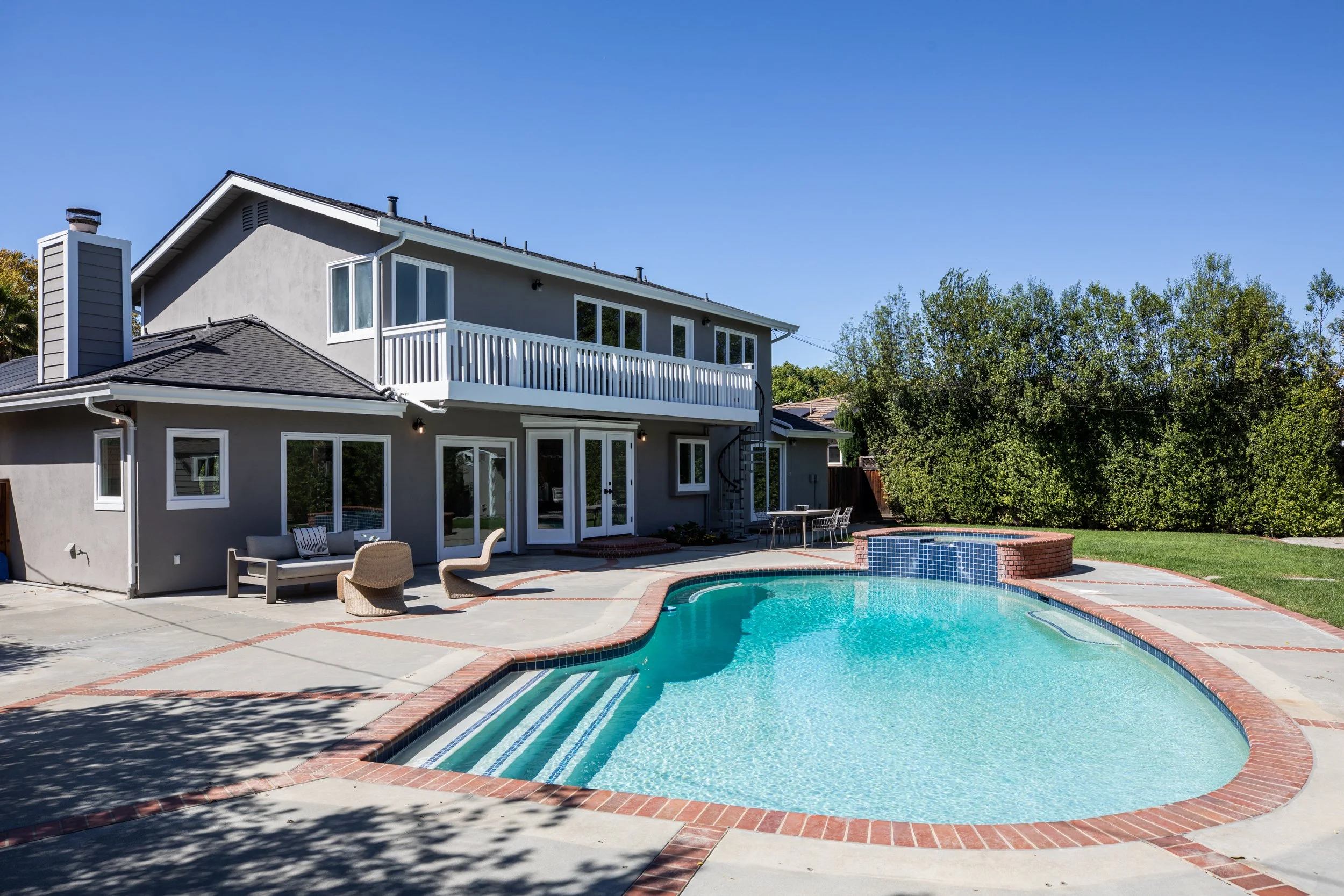 A backyard scene with a house featuring multiple windows, a second-floor balcony, and an outdoor area with a swimming pool surrounded by patio furniture and trees.