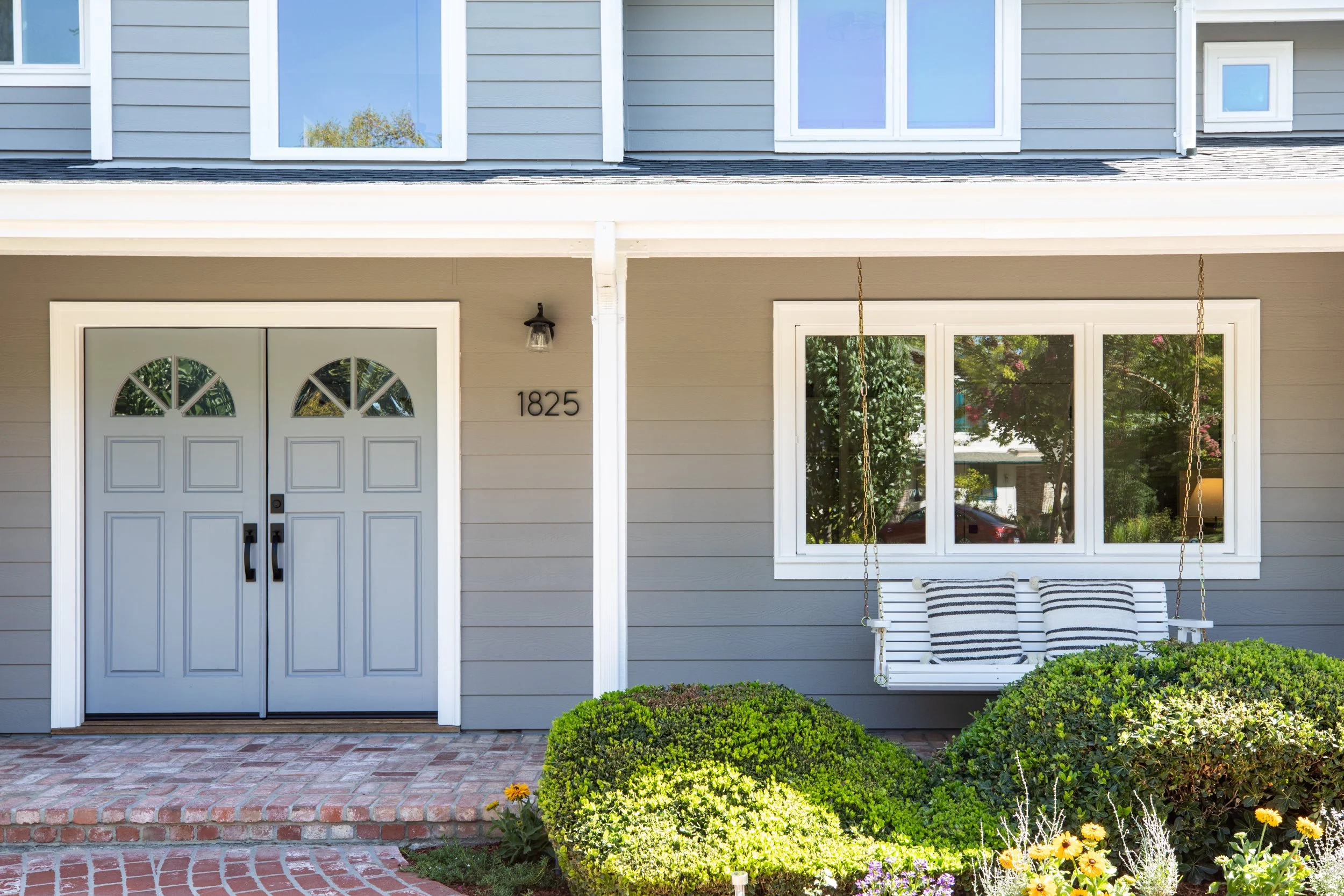 Front porch of a house with a double door, a window to the right, and a swing with black and white striped cushions. The house number is 1825. There are bushes and flowers in the garden.