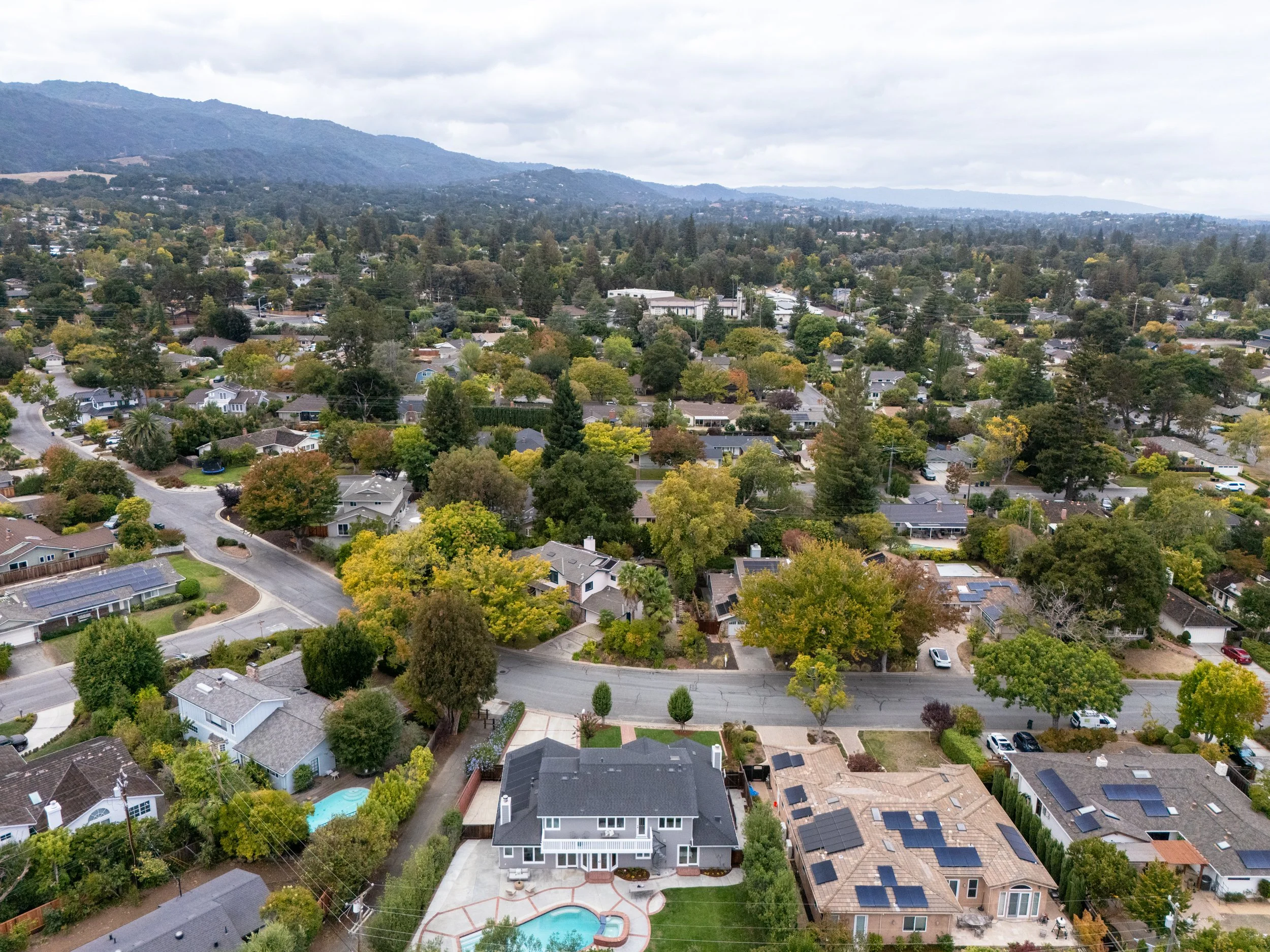 An aerial view of a suburban neighborhood with houses, trees, and roads, with mountains in the background under a cloudy sky.