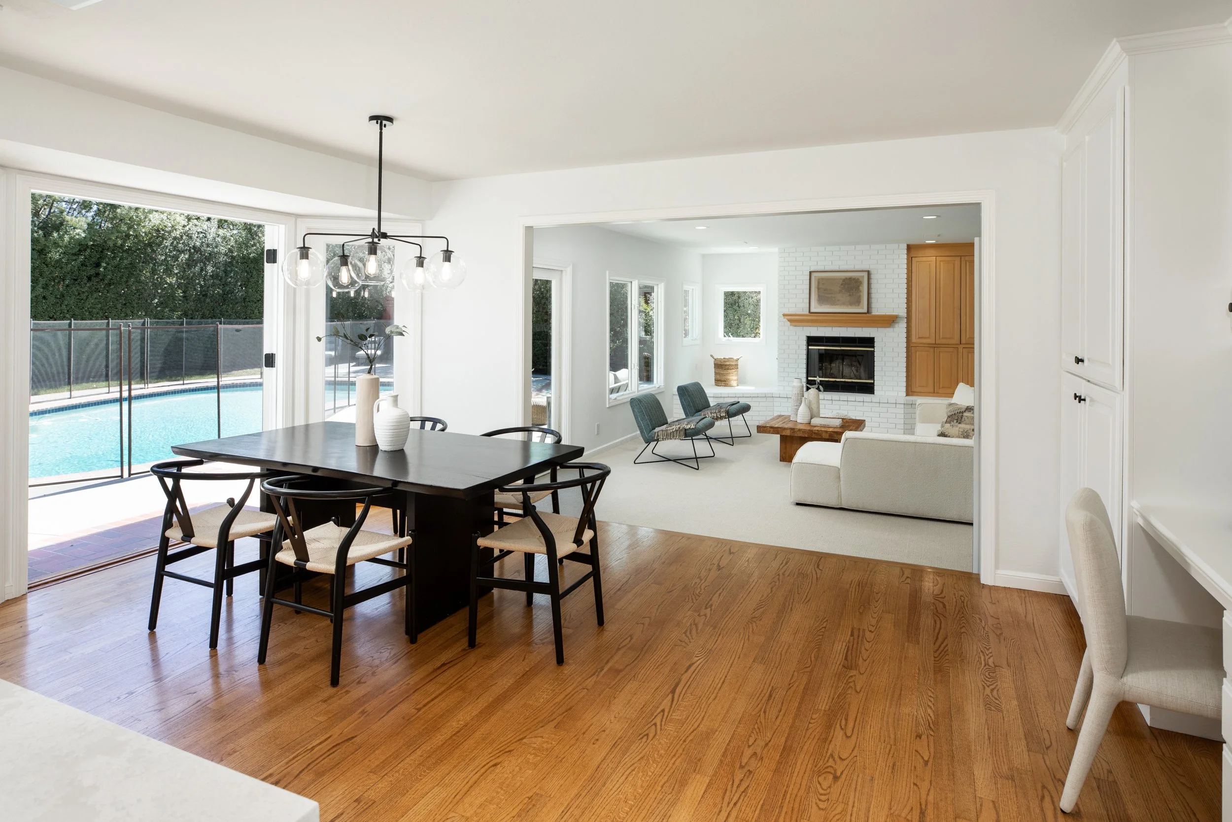 A bright, modern living room with a dining area next to large glass sliding doors leading to a backyard with a swimming pool. The room features white walls, a wooden floor, a black dining table with chairs, and a white couch with a fireplace and wind