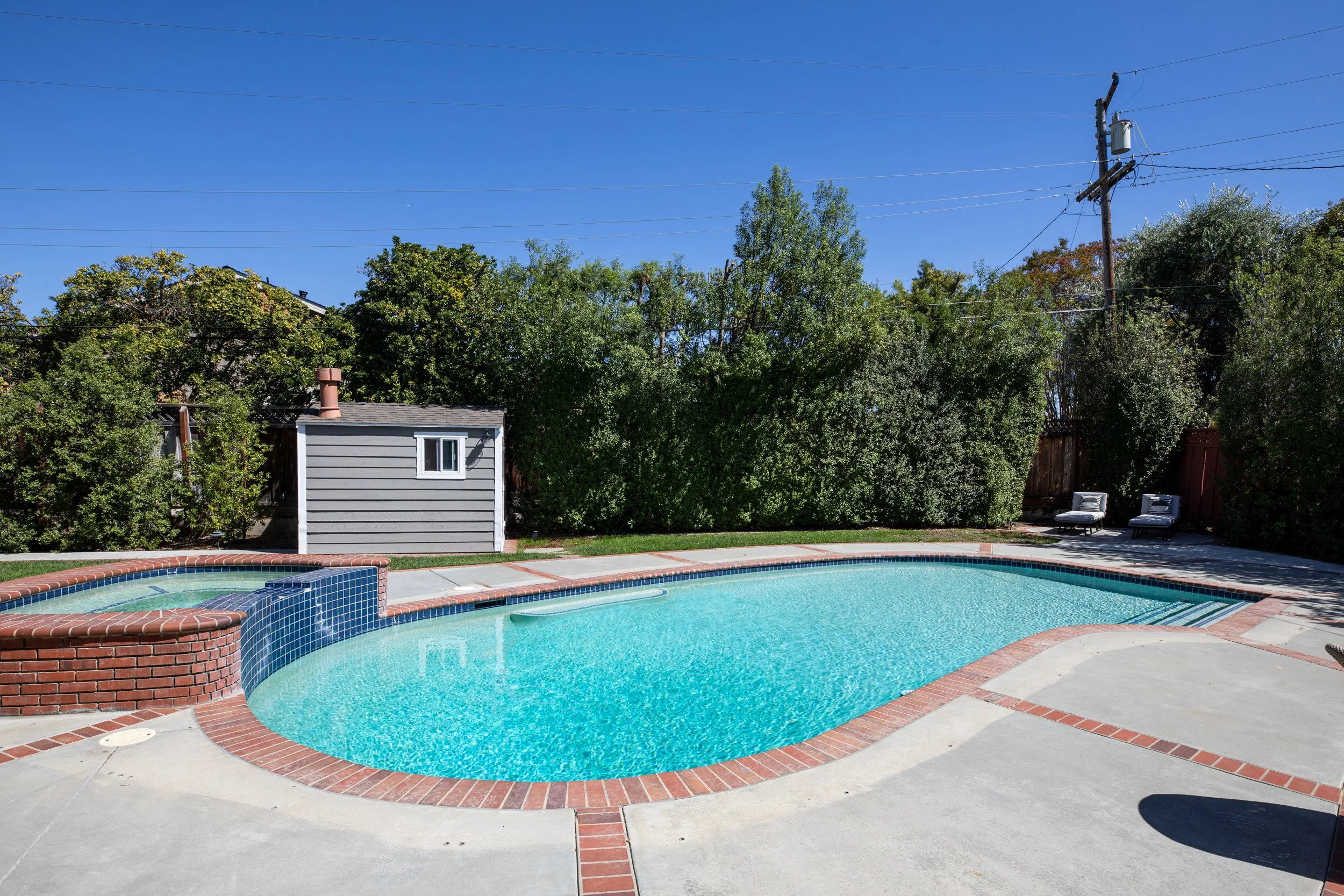 A backyard with a swimming pool, surrounded by a concrete patio with brick accents, and a background of trees, utility pole, and clear blue sky.