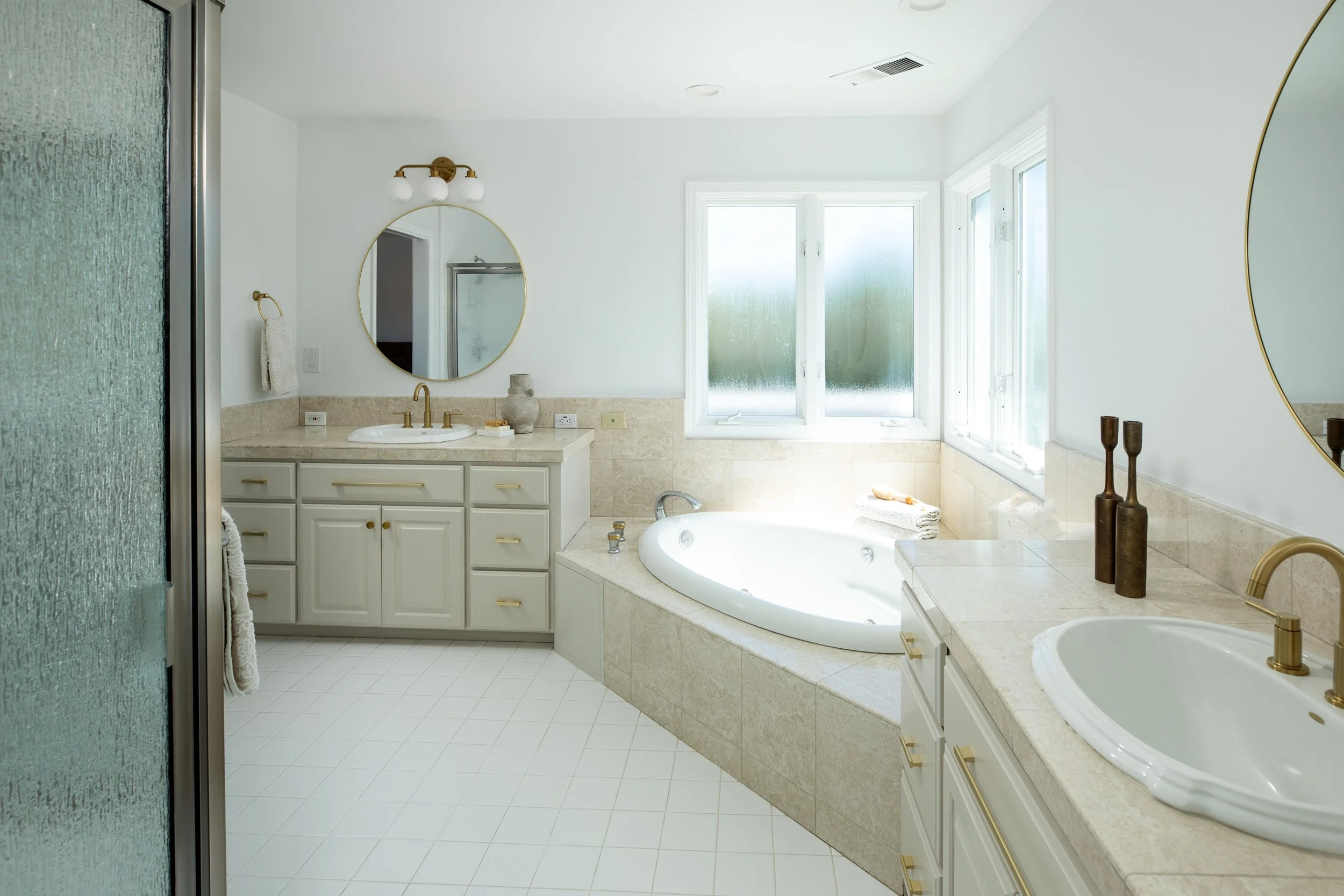A spacious bathroom with two oval sinks, a large bathtub, light-colored cabinetry, and large windows with frosted glass for privacy.