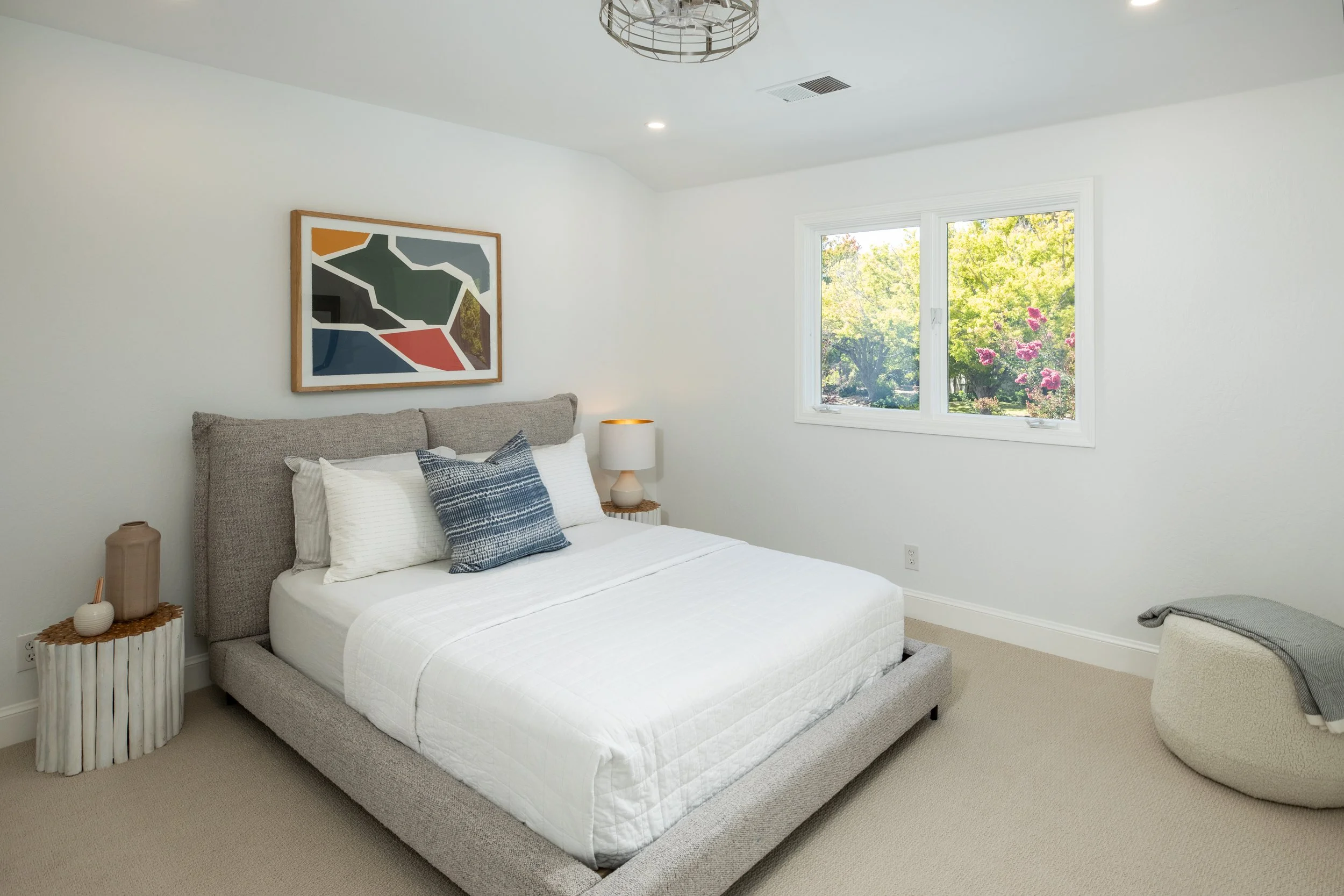 A bedroom with a bed, nightstand, and artwork on the wall. The bed has a striped pillow and white bedding. A window shows trees and pink flowers outside.