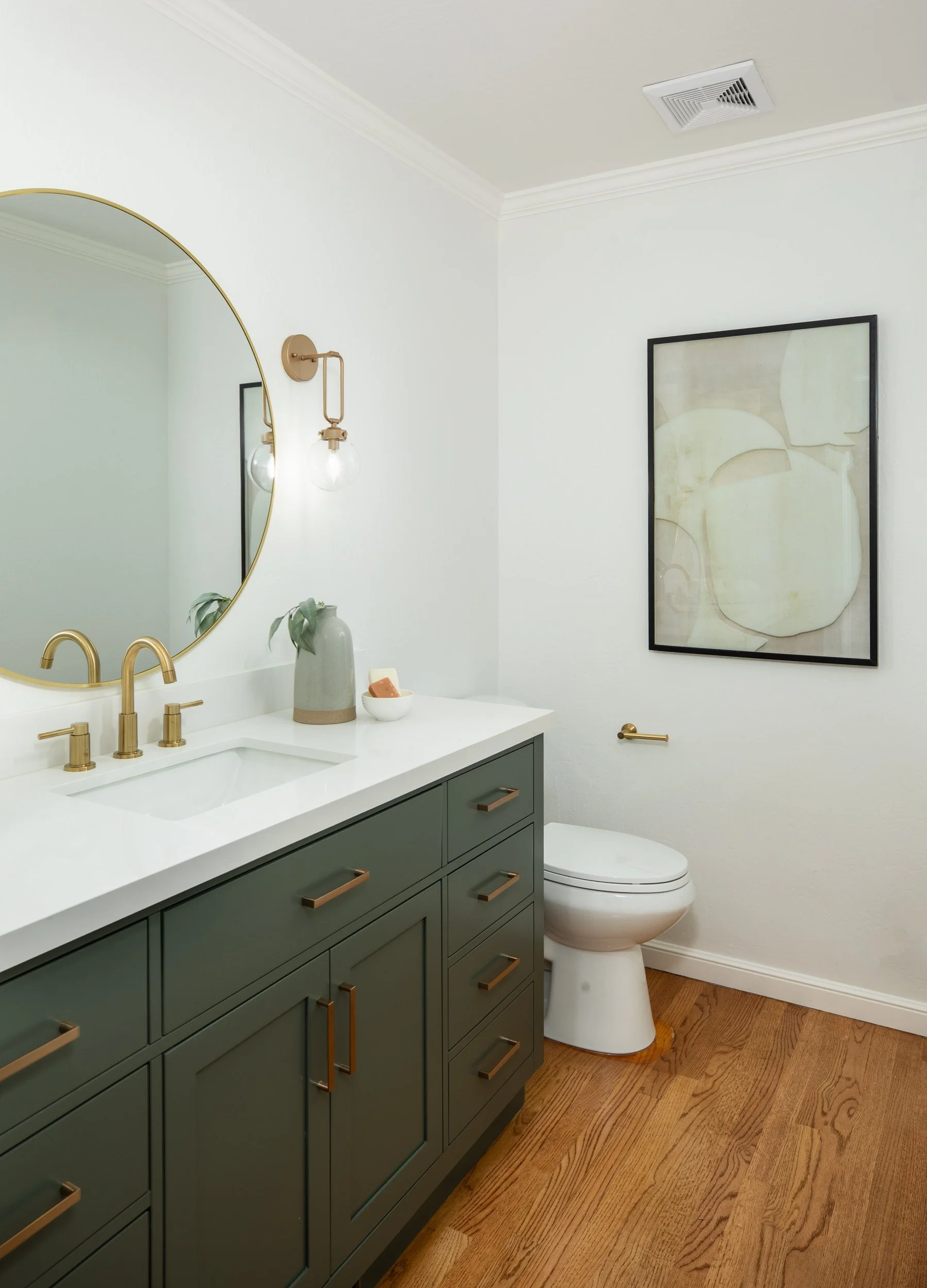 Modern bathroom with a green vanity, white countertop, gold fixtures, a round mirror, wall-mounted light, artwork on the wall, and a toilet.