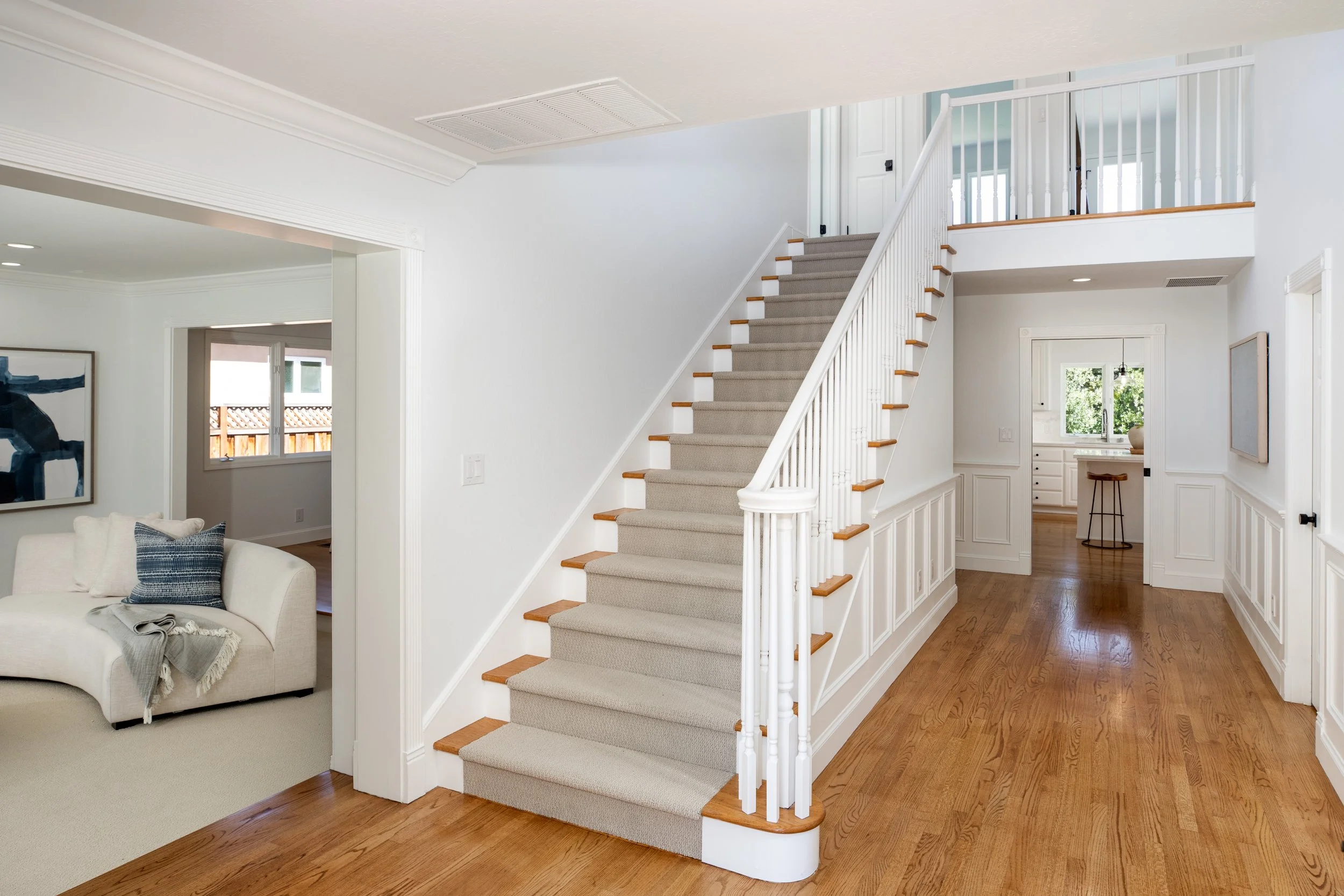 Interior of a home with a staircase with white railings and beige carpet, wooden floors, and white walls. Visible living room with a white sofa and pillows, a side table, and artwork on the walls. Kitchen area visible in the background.