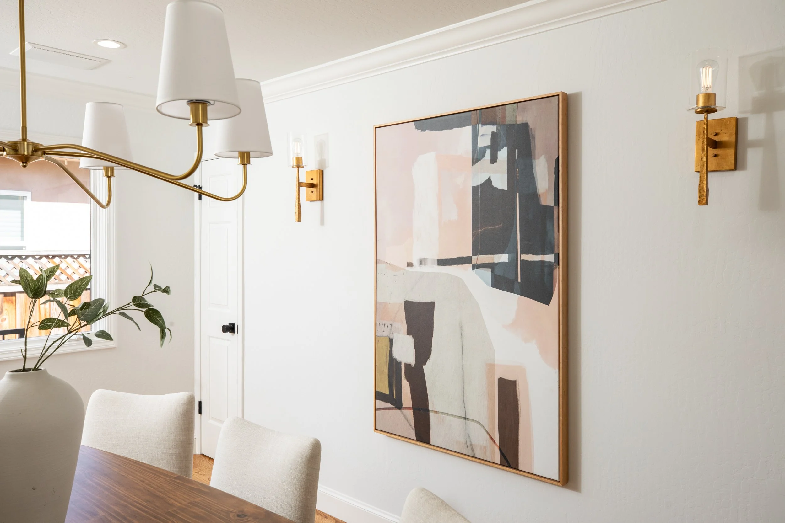 A dining room with a wooden table, white upholstered chairs, a large abstract painting on the wall, a potted plant, and three light fixtures including a chandelier and wall sconces.