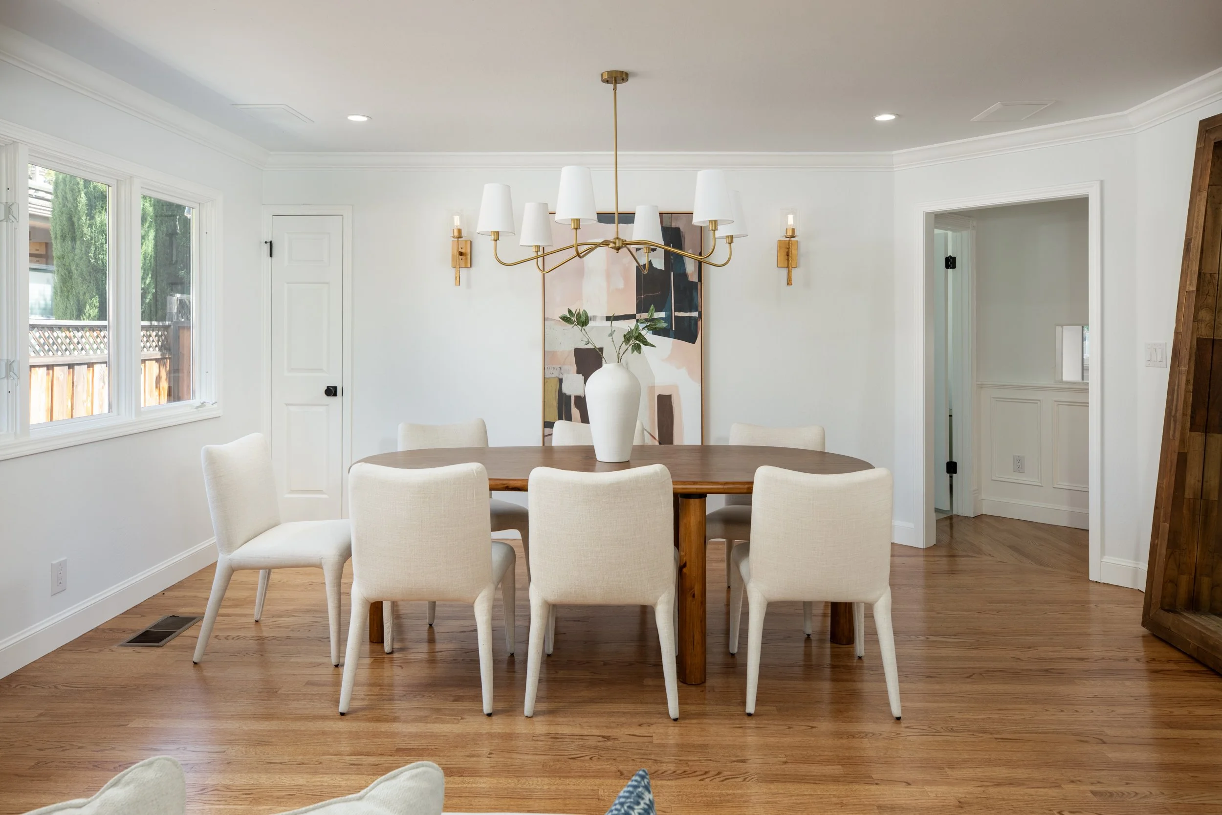 A dining room with a wooden table surrounded by white upholstered chairs. A large white vase with greenery is in the center of the table. A modern chandelier with white shades hangs above the table. The room features white walls, crown molding, and h