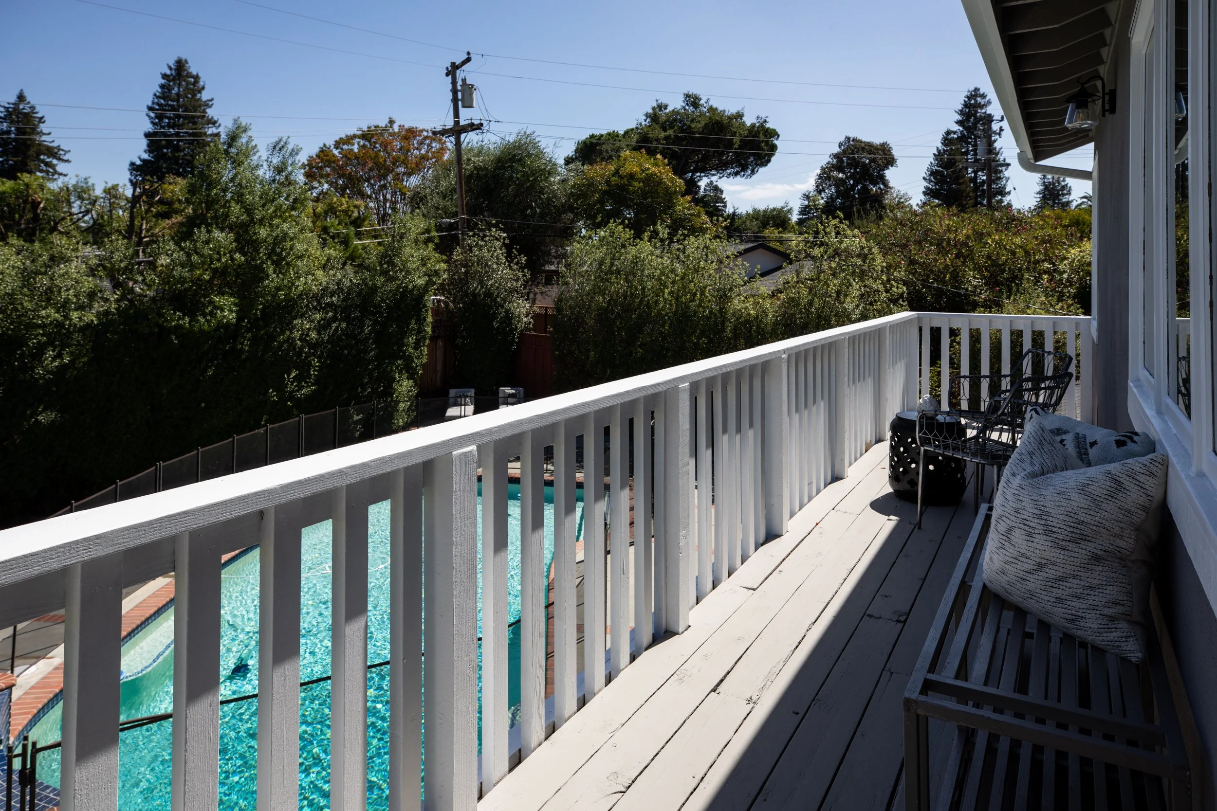 View from a balcony with white railing overlooking a swimming pool, surrounded by green trees and clear blue sky.