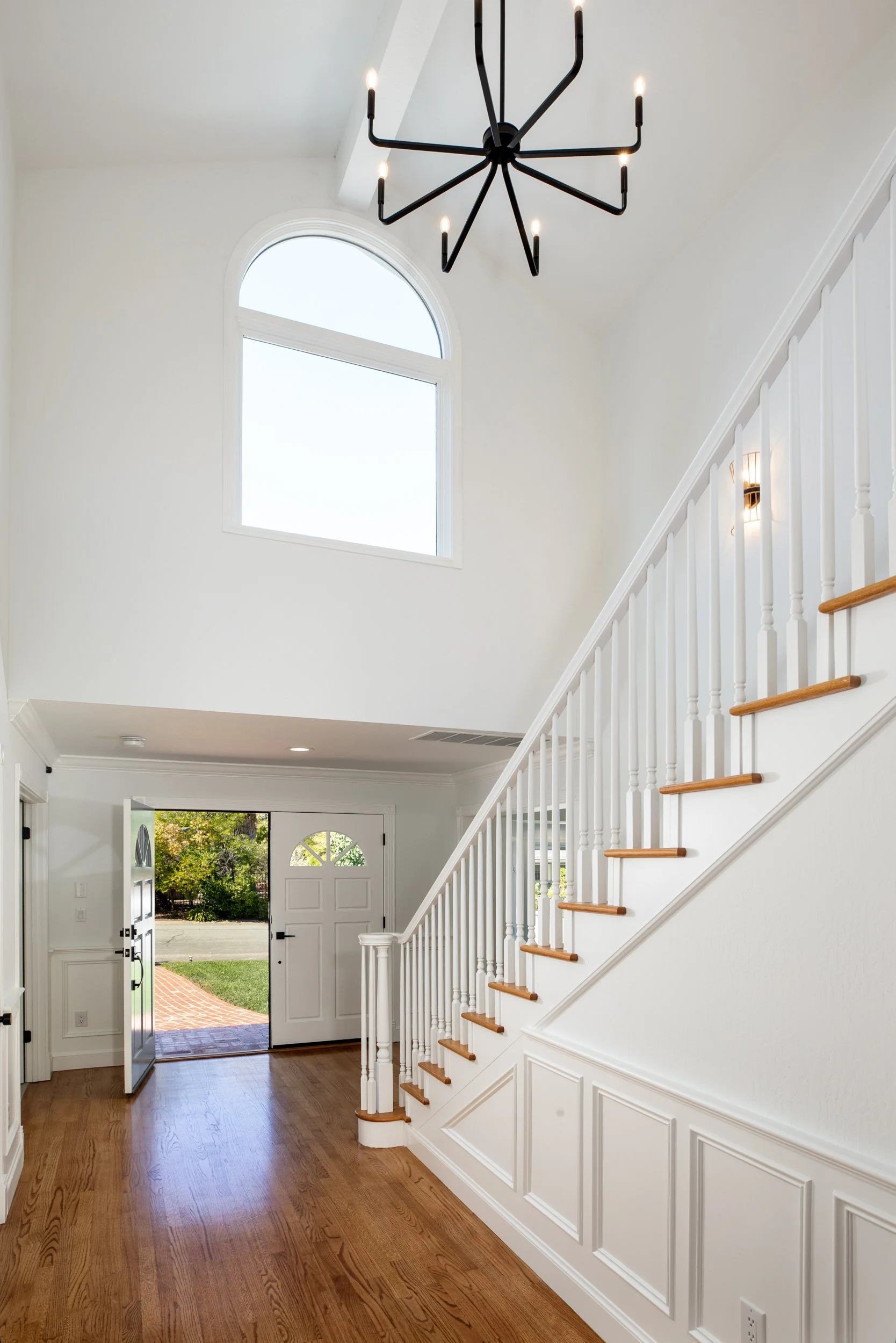 Entryway of a house with a staircase, large front door open to outdoors, and a window above the staircase. Modern chandelier hanging from ceiling, hardwood floors, white walls, and wall sconce lighting.
