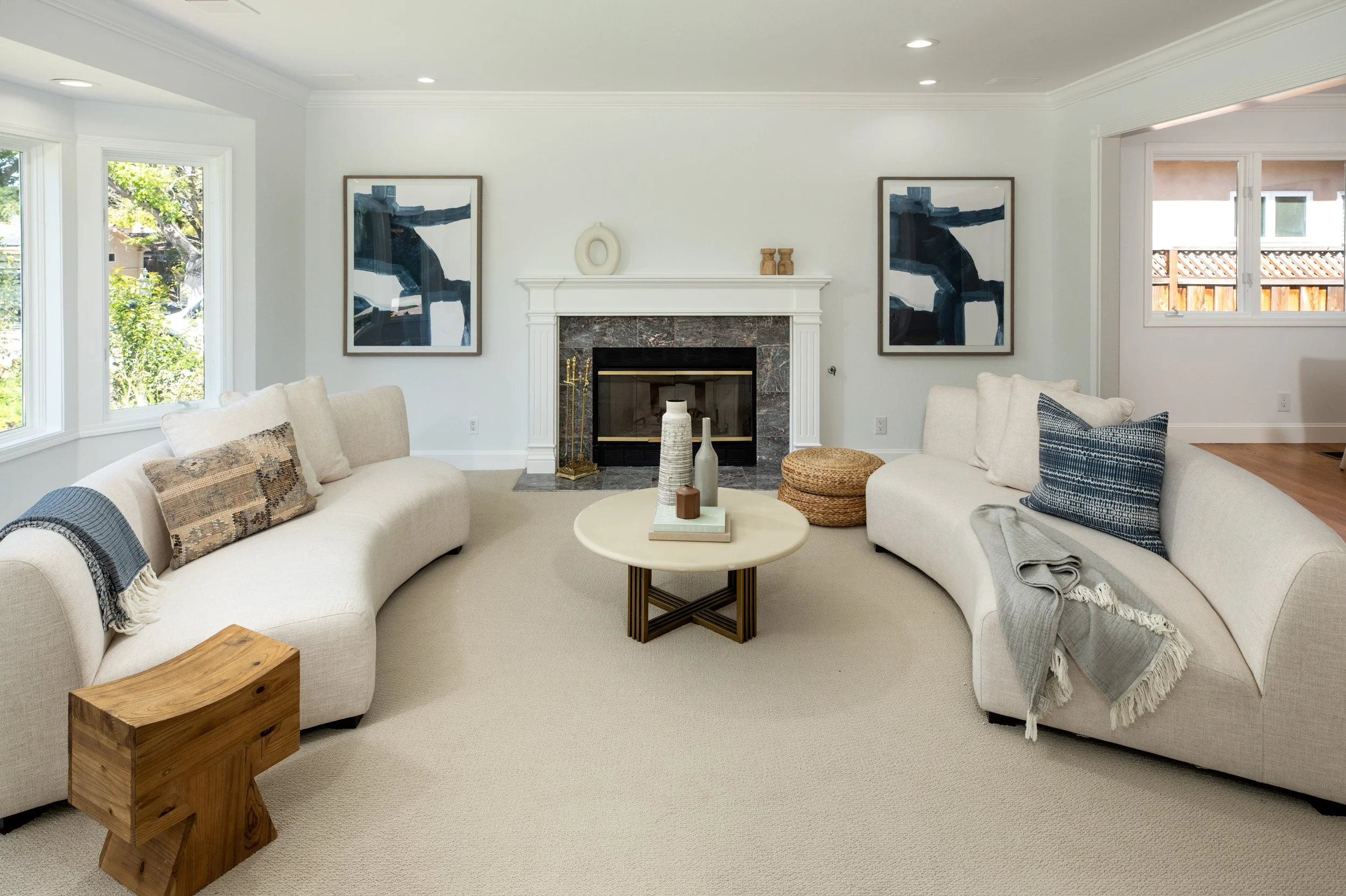 A living room with two cream-colored sofas, decorated with pillows and throws. There is a round coffee table with decorative vases, a wooden stool, and a fireplace with artwork and decorative objects on the mantel. Large windows offer a view of a bac