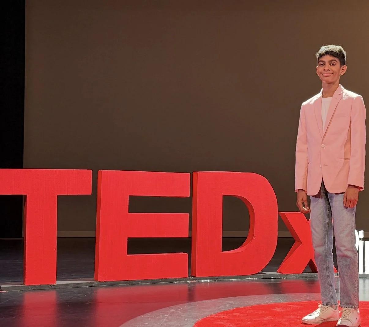 A young man in a pink blazer and jeans standing on a TEDx stage with red TEDx letters and a dark background.