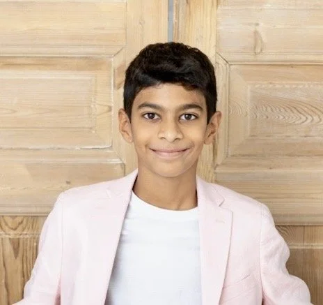 A young boy with dark hair, wearing a light pink blazer and white shirt, smiling at the camera in front of a wooden panel background.