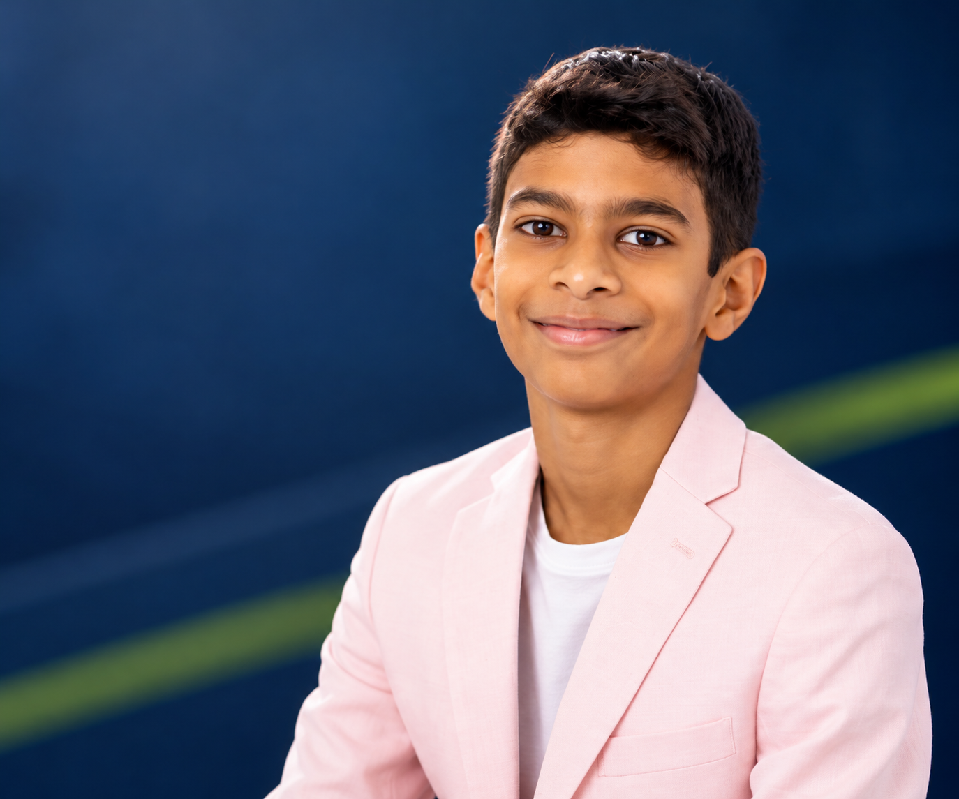 A young boy with short dark hair, wearing a light pink blazer over a white shirt, smiling at the camera against a blurred blue and green background.