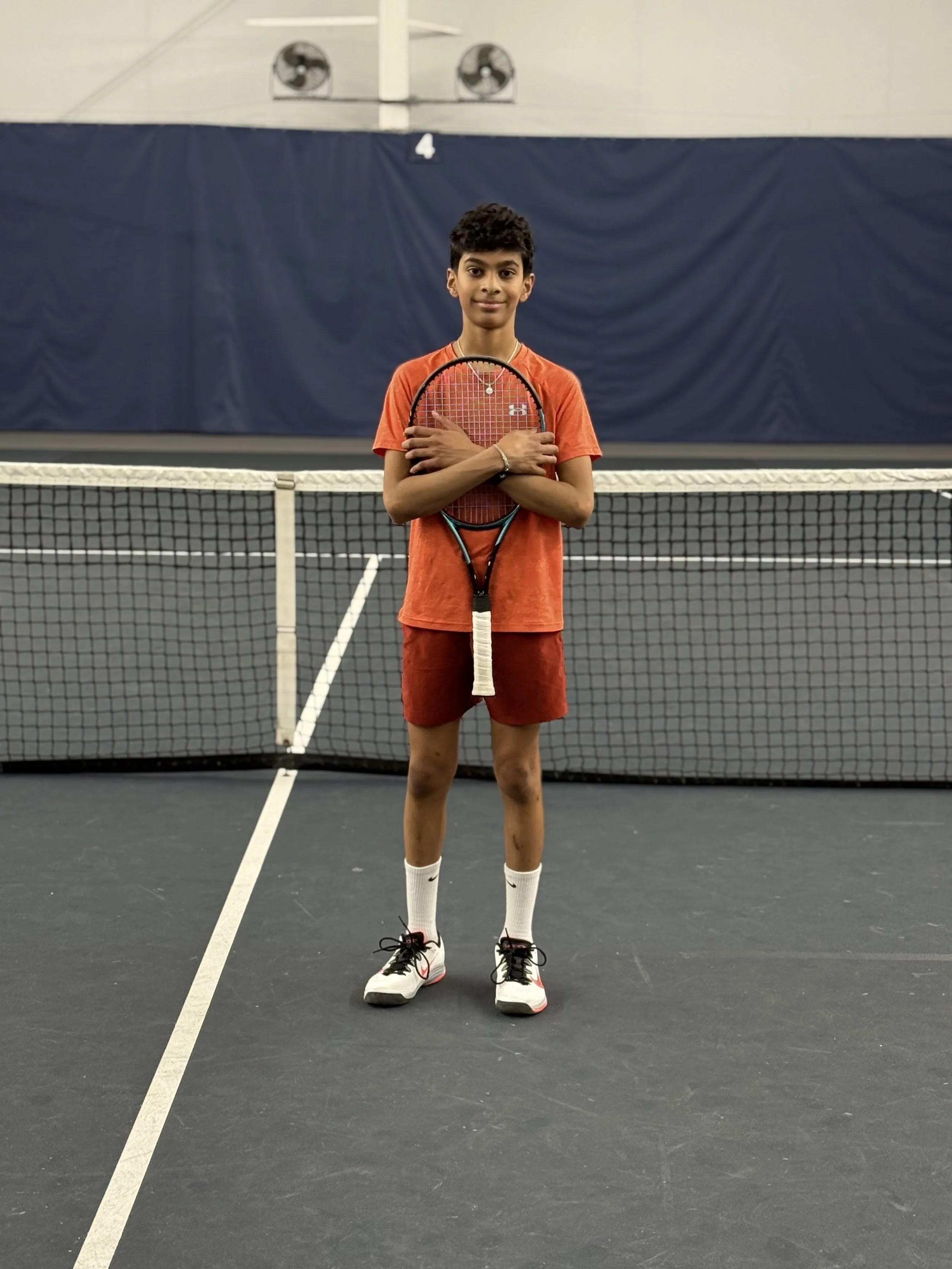 A young boy standing on an indoor tennis court, holding a tennis racket in front of his chest with crossed arms. He is wearing a red T-shirt, red shorts, white socks, and white sneakers. There is a tennis net in front of him and a blue curtain with a number 4 hanging on it in the background.
