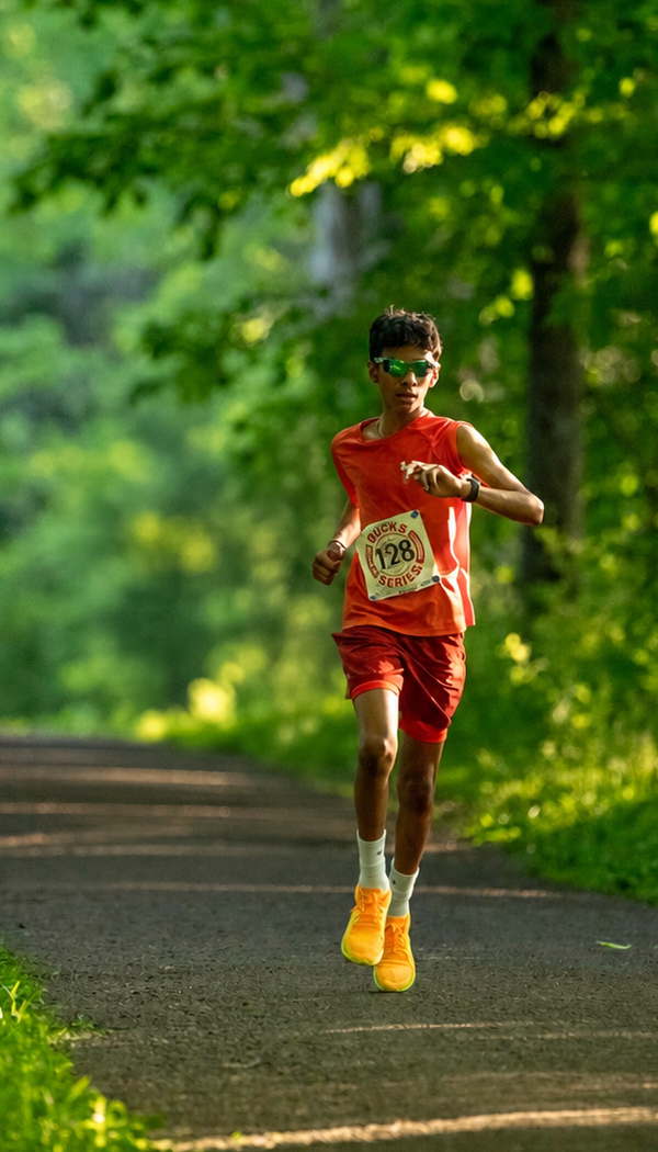 Young male runner wearing sunglasses, an orange athletic outfit, and yellow running shoes, racing on a forest trail during daytime.