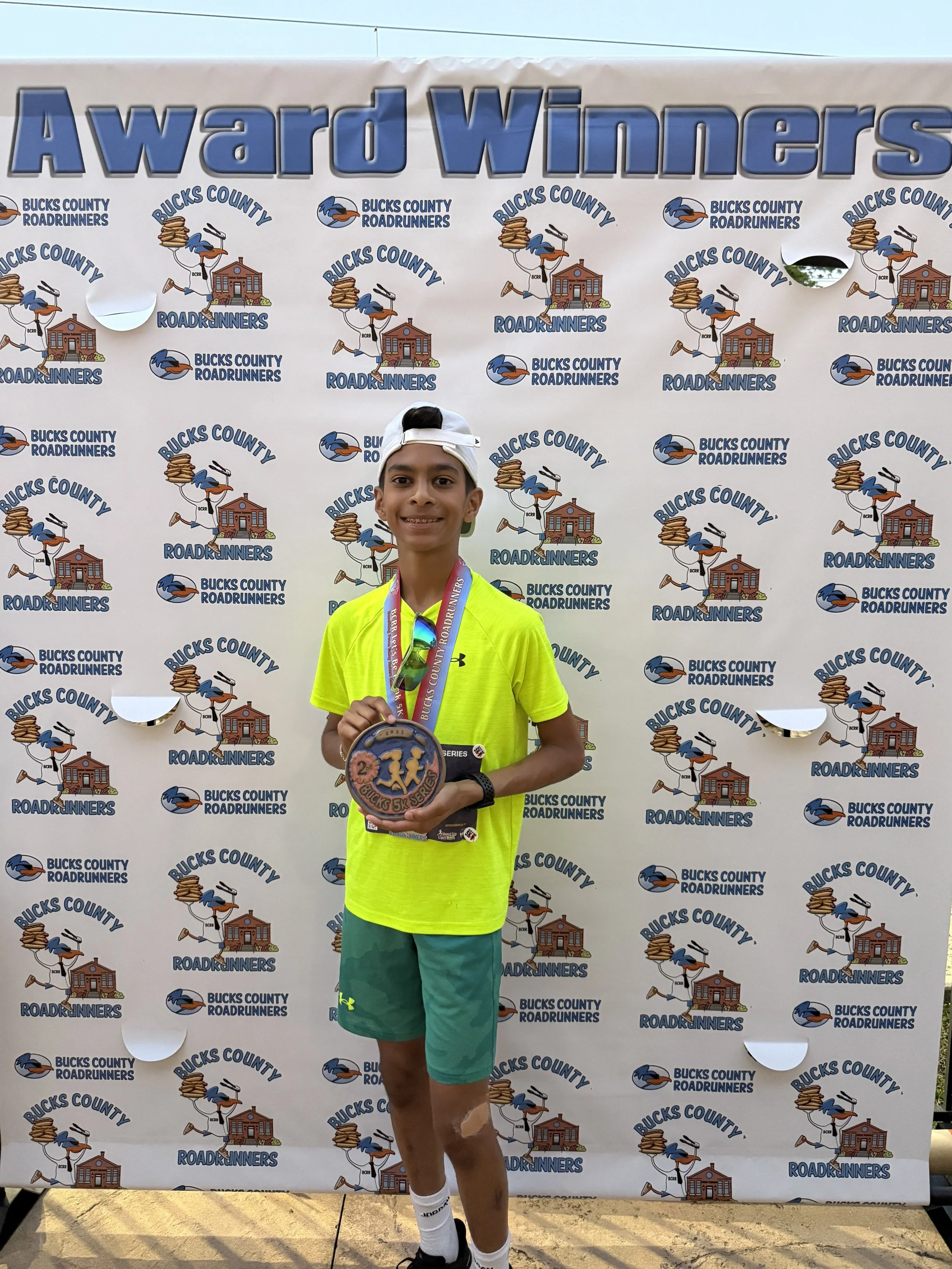 Young boy holding a medal and standing in front of a backdrop with the text "Award Winners" and logos for Bucks County Roadrunners, featuring a running bird and a building.