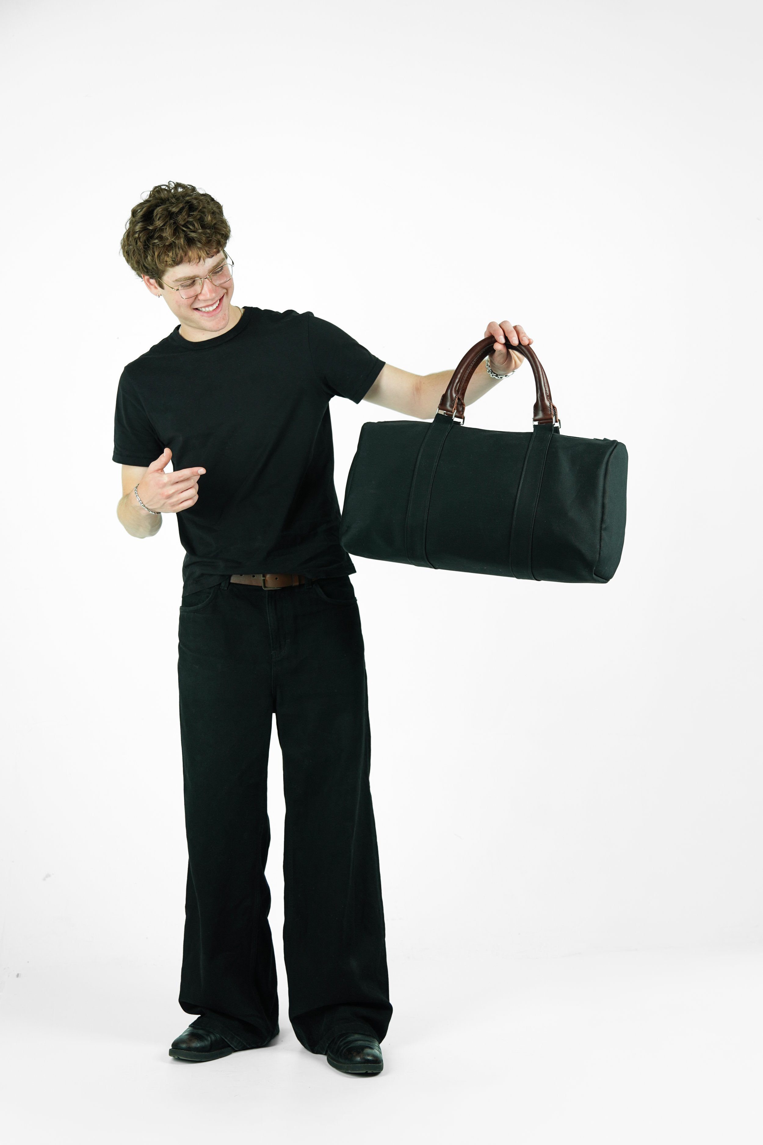 A young man with curly hair, glasses, wearing a black shirt and black pants, smiling and pointing at a large black duffel bag with brown handles, standing against a white background.