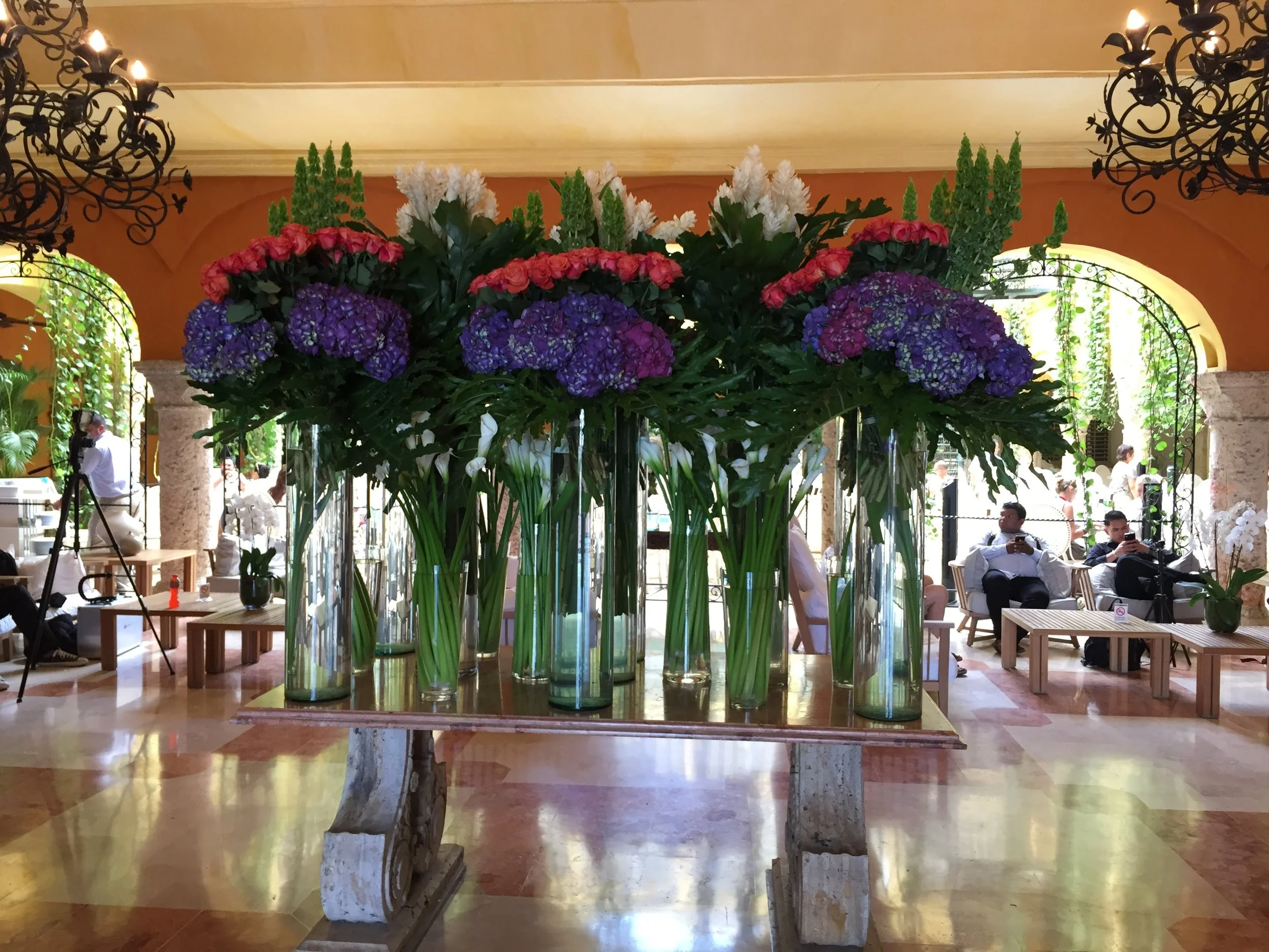 A large floral arrangement featuring purple hydrangeas, pink roses, and white flowers in tall glass vases on a wooden table, inside a spacious room with high ceilings, chandeliers, and people sitting and standing around.