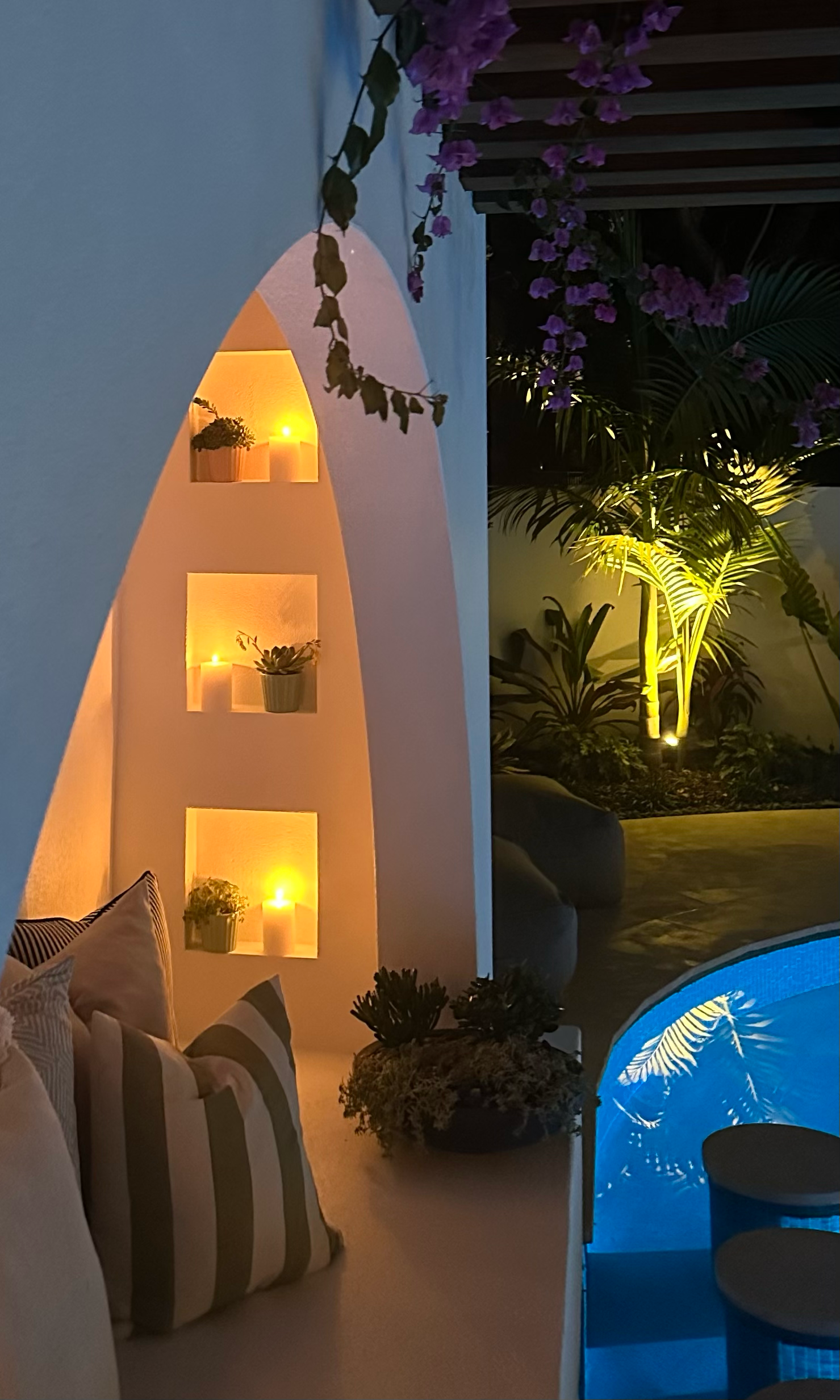 Outdoor pavilion scene at night with illuminated built-in wall shelves with candles and plants, purple flowering vine hanging, and lush green plants lit by landscape lighting.