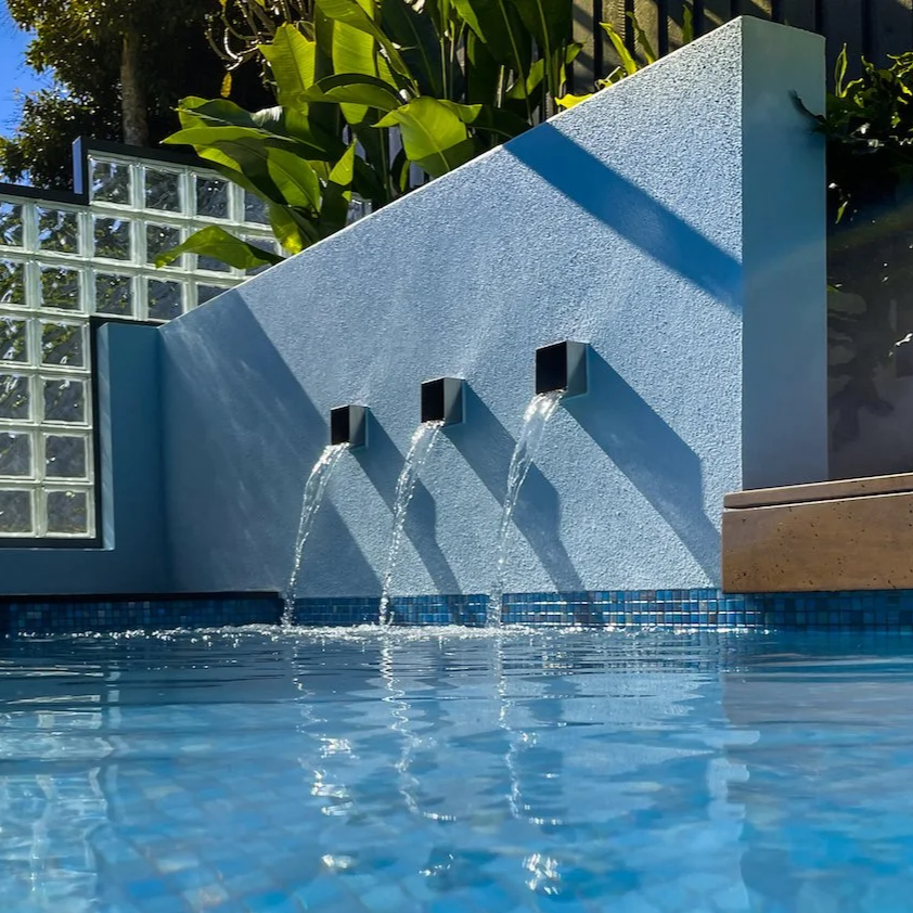 Three black wall-mounted spouts pouring water into a swimming pool with blue tiles, lush green plants, and a glass block wall in the background.