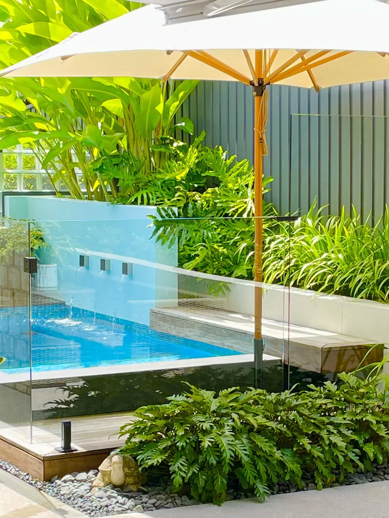 A backyard scene with a swimming pool, surrounded by lush green plants and a large beige patio umbrella providing shade, with a gray fence in the background.