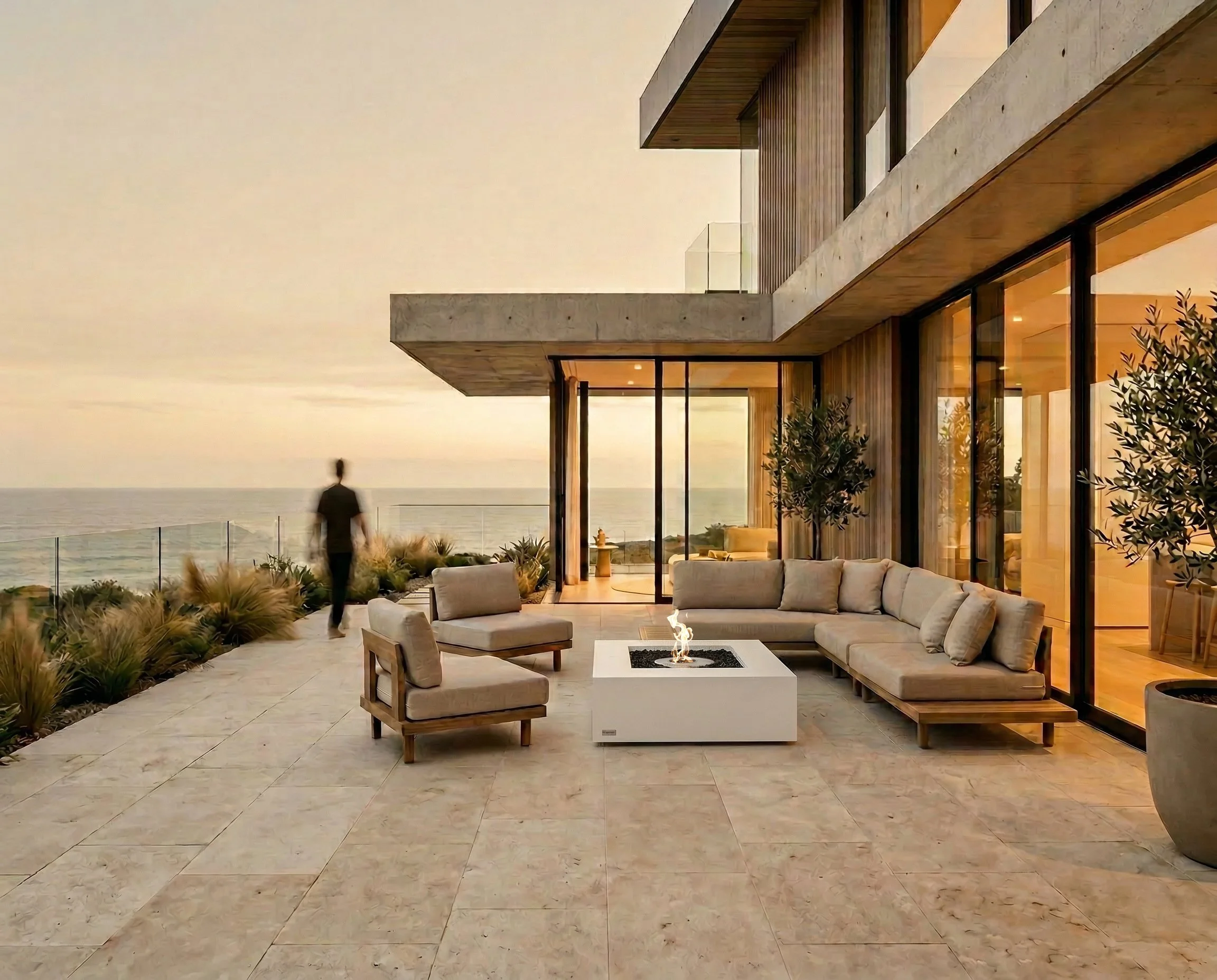 Modern house with large glass windows and a balcony overlooking the ocean at sunset, outdoor seating area with beige cushions and a fire pit, plants and greenery around.