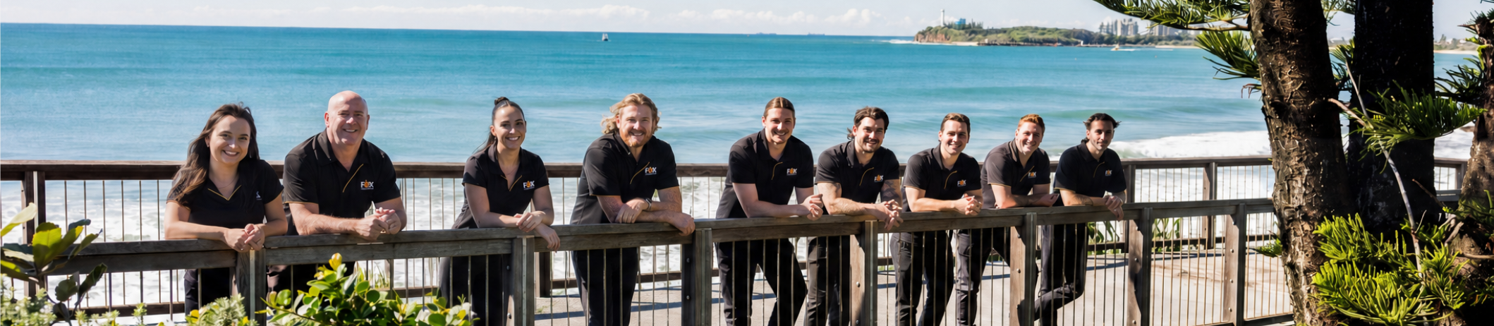 Group of nine people standing behind a wooden railing by the beach with ocean and sky in the background.