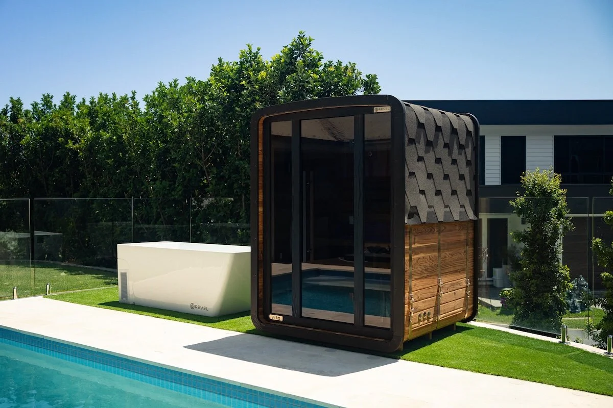 Modern outdoor poolside cabana with glass sliding doors, wooden walls, shingle roof, next to a swimming pool, with greenery and a house in the background.