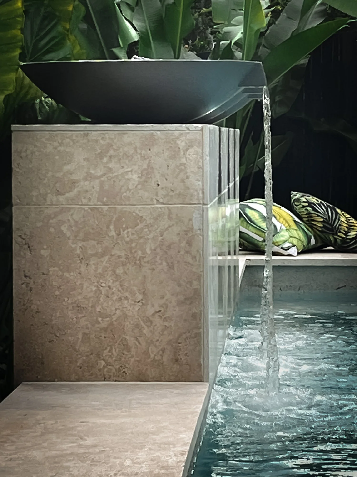 An outdoor water fountain with a tiered design, water flowing from the top tier into a pool below, surrounded by lush green plants.