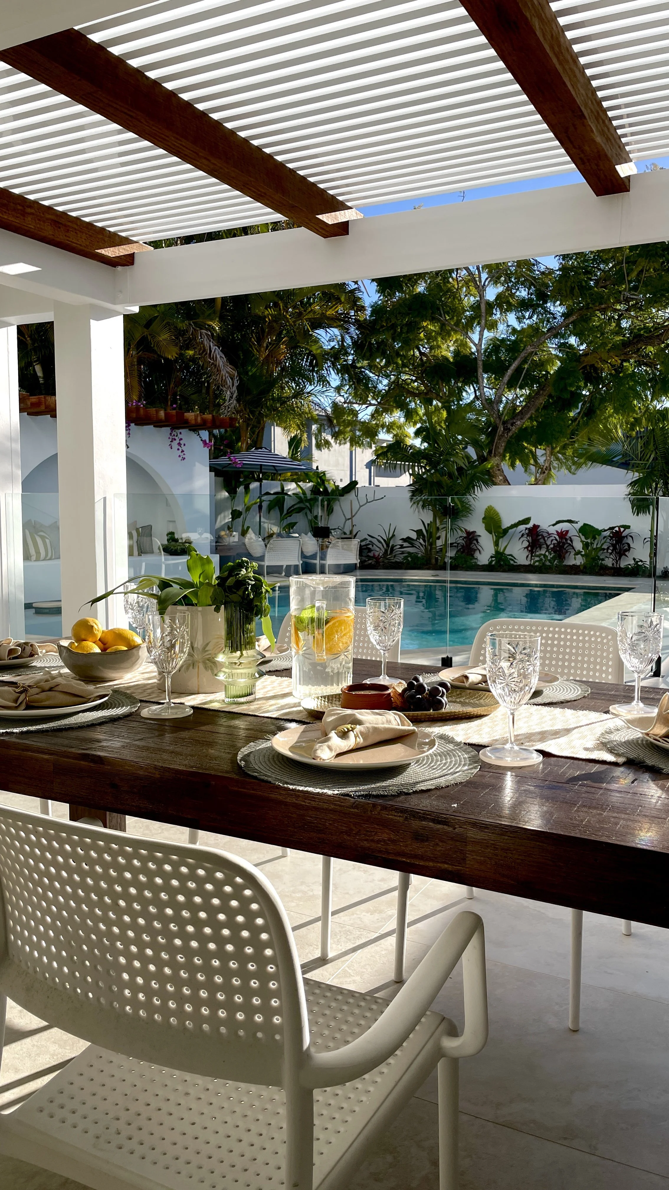 A dining table set with plates, glasses, napkins, and a pitcher of water with lemon slices next to a swimming pool.