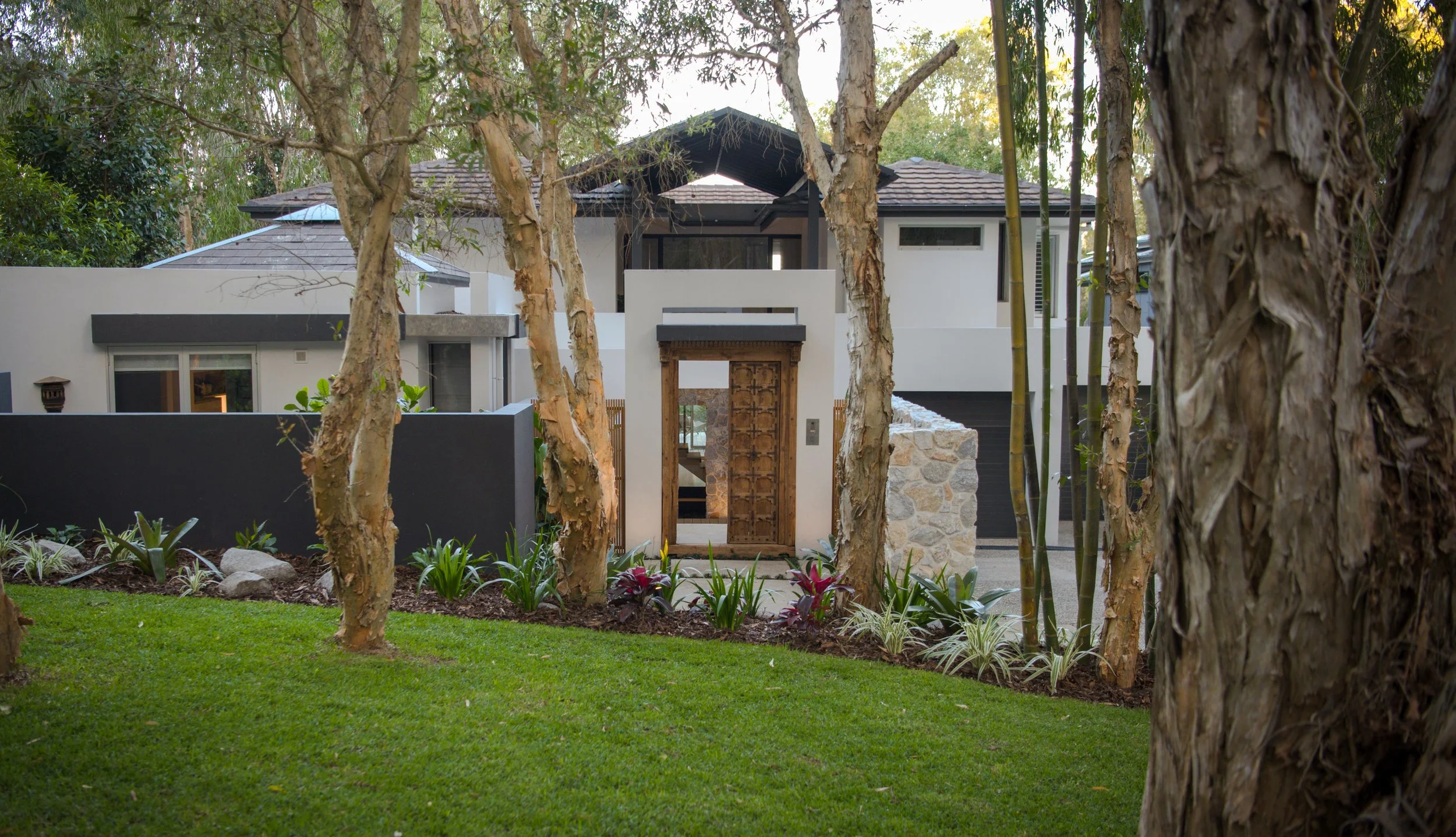 Front view of a modern house partly hidden behind trees and a white wall, with a wooden door and greenery in the front yard.
