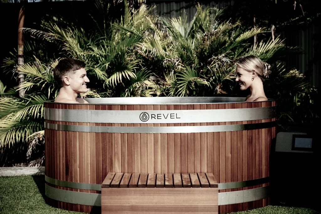 Two people smiling and relaxing in an ice bath labeled 'Revel' with lush green plants in the background.