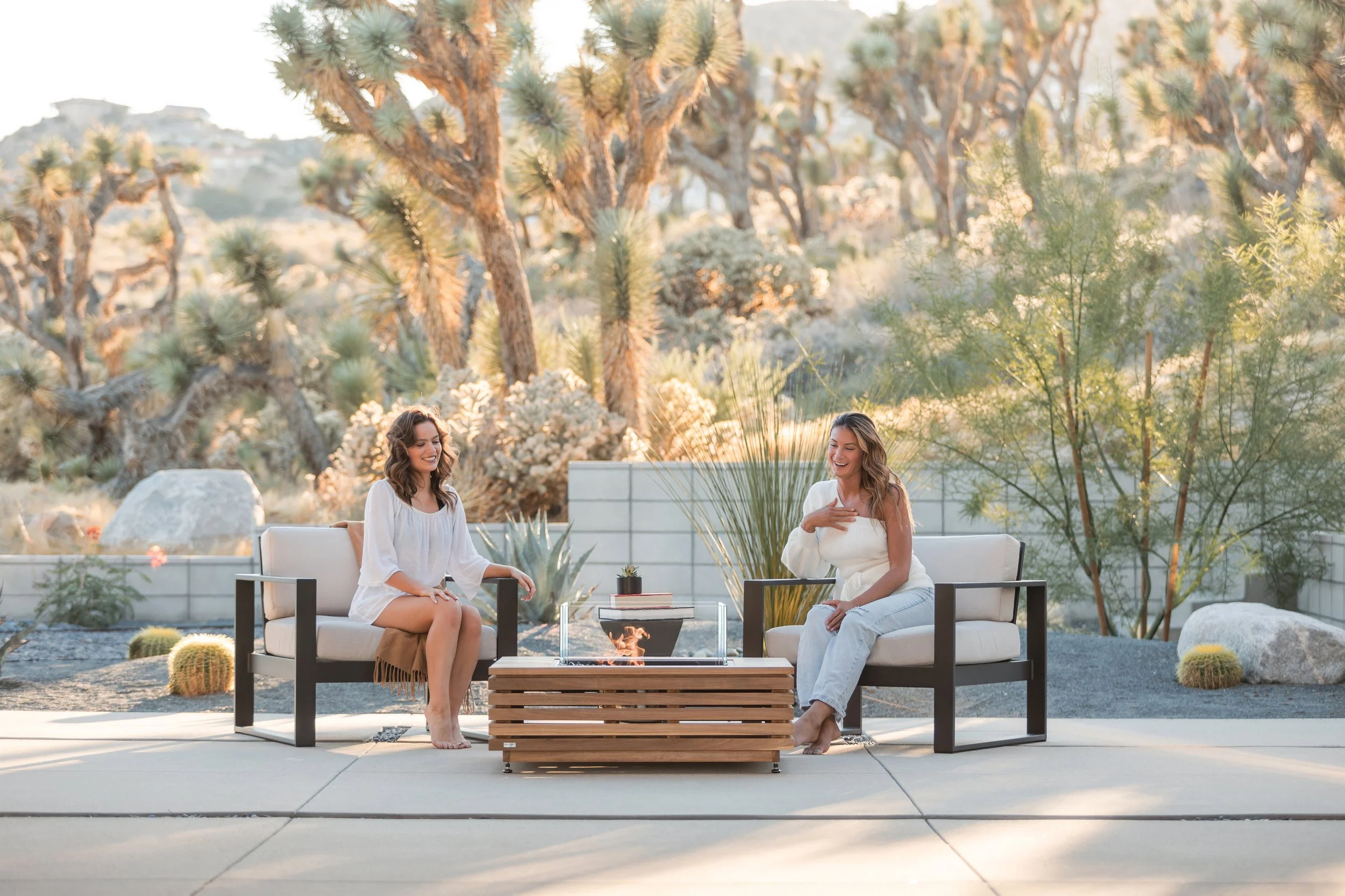 Two women sitting outdoors on patio furniture, smiling and talking, with a desert landscape featuring tall Joshua trees in the background during sunset