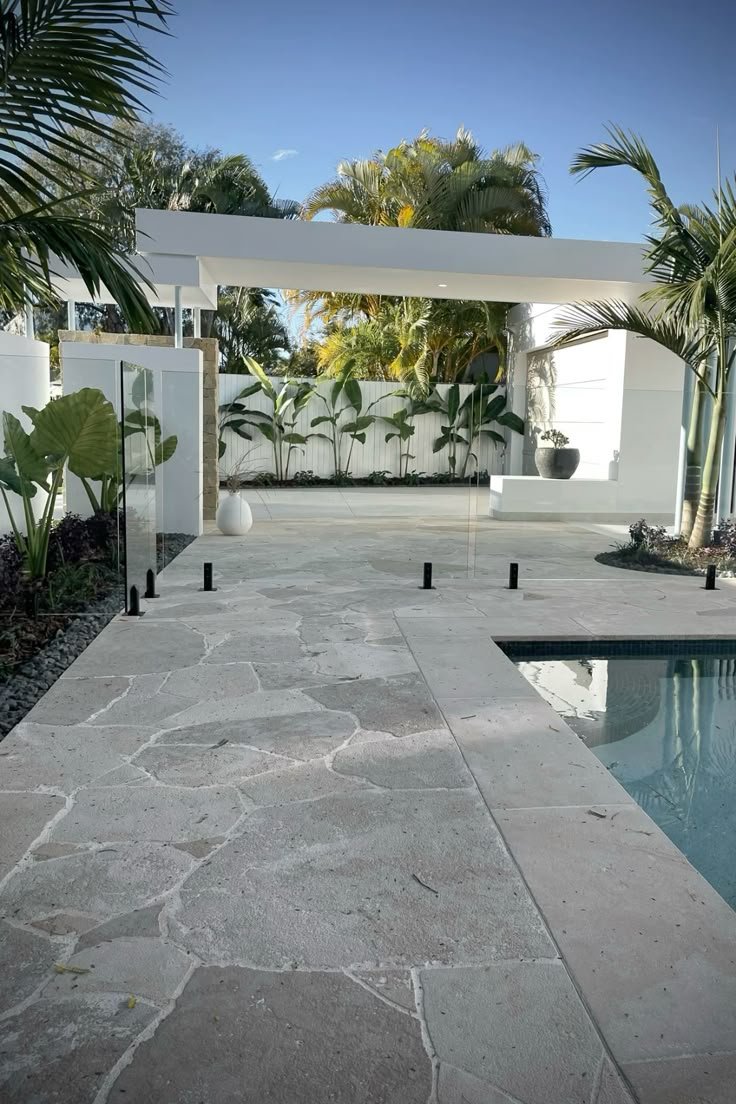 Modern backyard with a stone pathway, swimming pool, tropical plants, and a white minimalist structure under a partly cloudy sky.