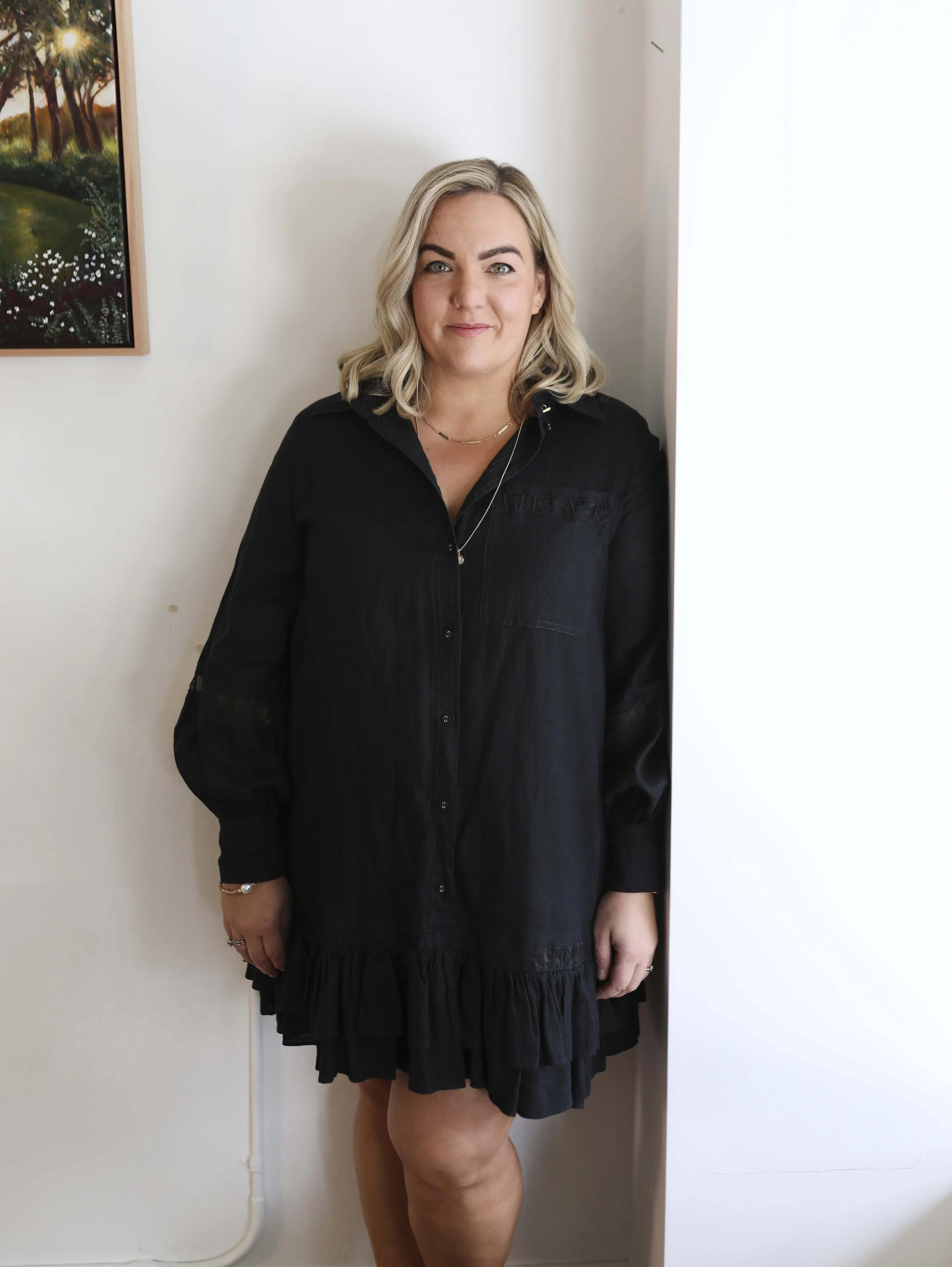 A woman with blonde wavy hair, wearing a black dress and jewelry, standing indoors near a white wall and a partially visible painting.