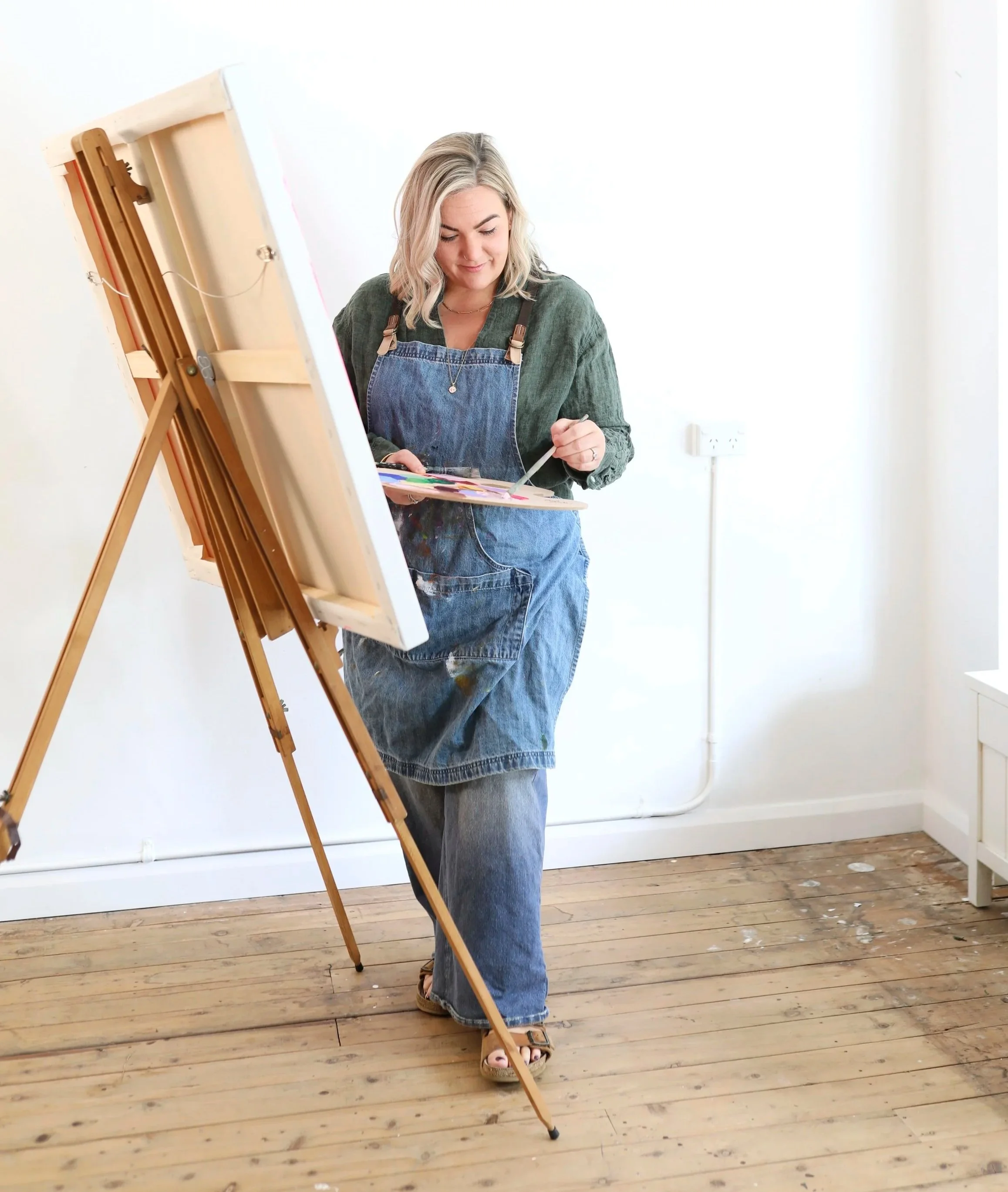A woman with shoulder-length blonde hair wearing a green shirt and blue overalls is painting on a canvas placed on an easel in a room with white walls and wooden flooring. She is holding a paintbrush in her right hand and a palette in her left hand, and is looking down at her work.
