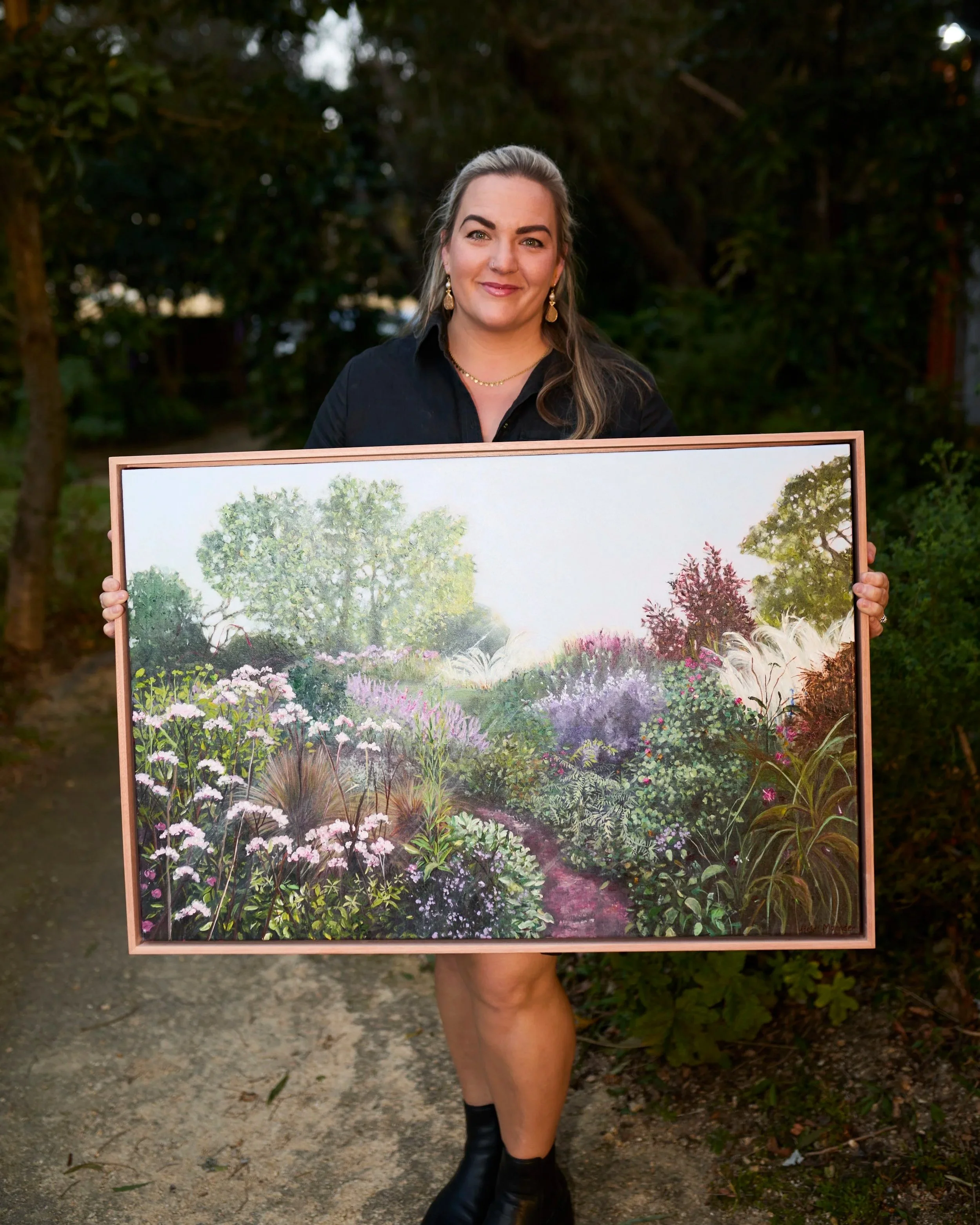 A woman with blonde hair and earrings holds a large landscape painting of trees and flowers outdoors. She wears a black shirt and black boots, standing on a dirt path with greenery in the background.