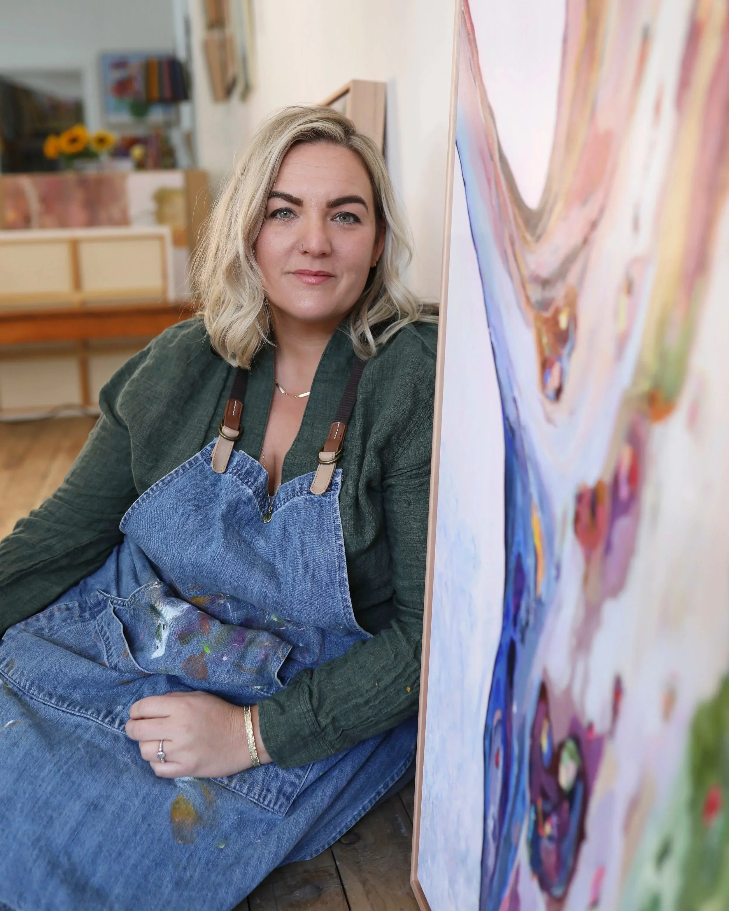 Heidi Maunder, sitting in an art gallery with her original colourful paintings on the walls.