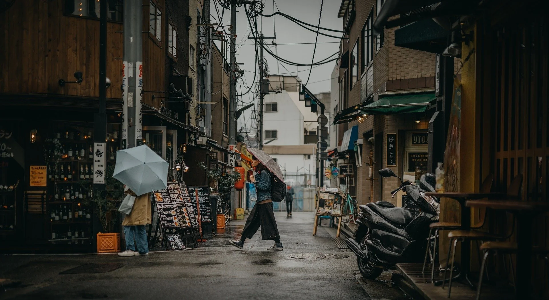A rainy street scene in Japan with two women holding umbrellas, one in a yellow jacket and the other in a denim jacket. The street is lined with small shops and restaurants, and there are motorcycles parked on the side. The sky is gray and the pavement is wet.