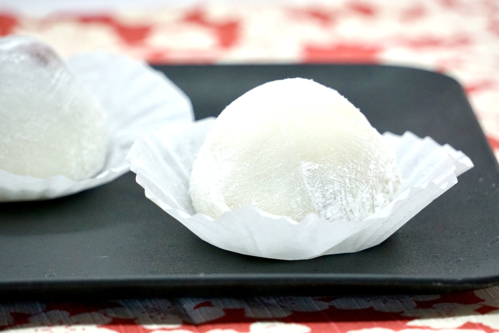 Two white, spherical mochi desserts in paper cups, placed on a black tray. The background shows a red and white patterned cloth.