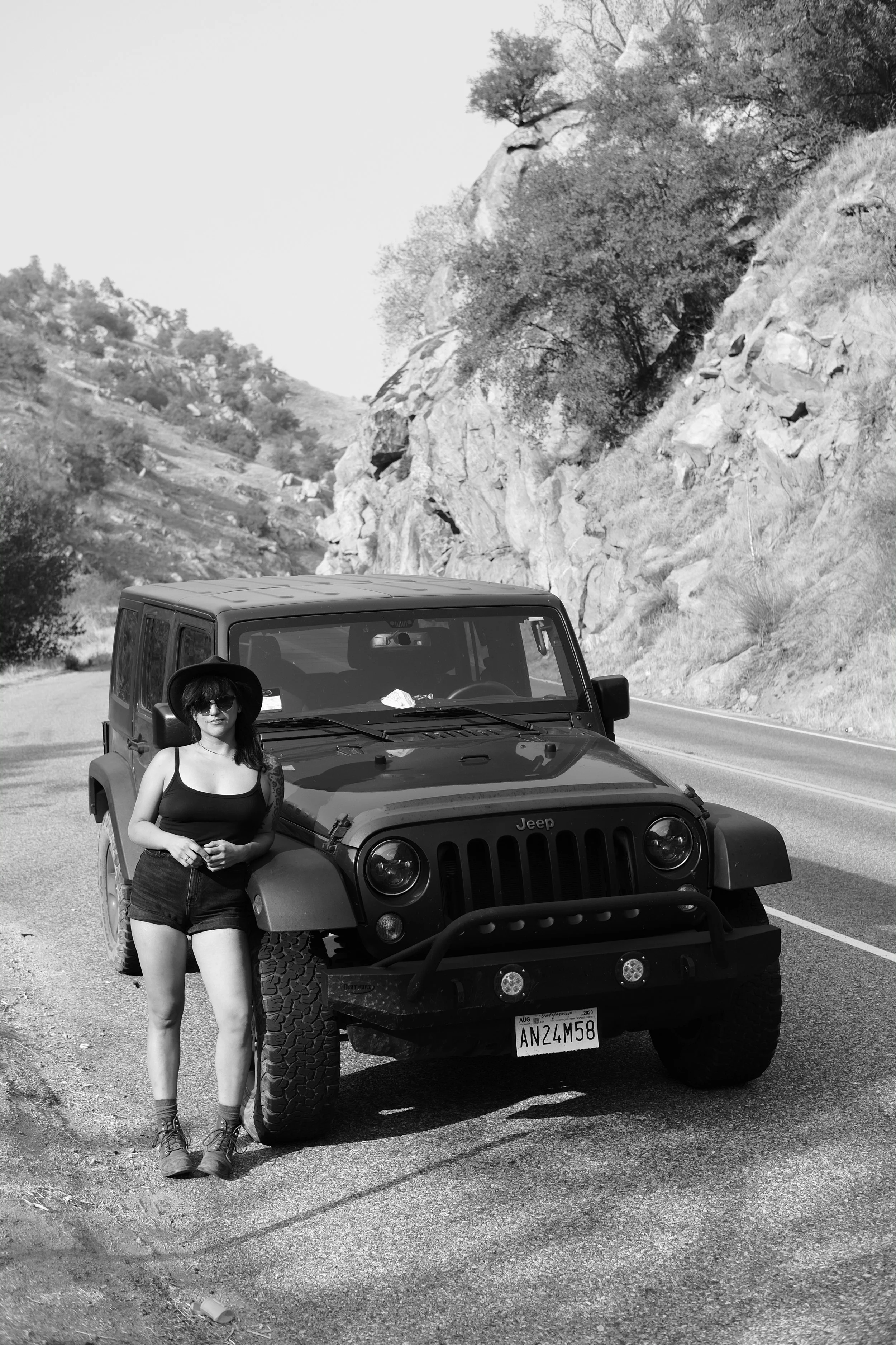A woman with tattoos, wearing sunglasses, a hat, a tank top, shorts, and boots, standing in front of a black Jeep SUV with California license plate, on a mountain road with rocky terrain and trees in the background.