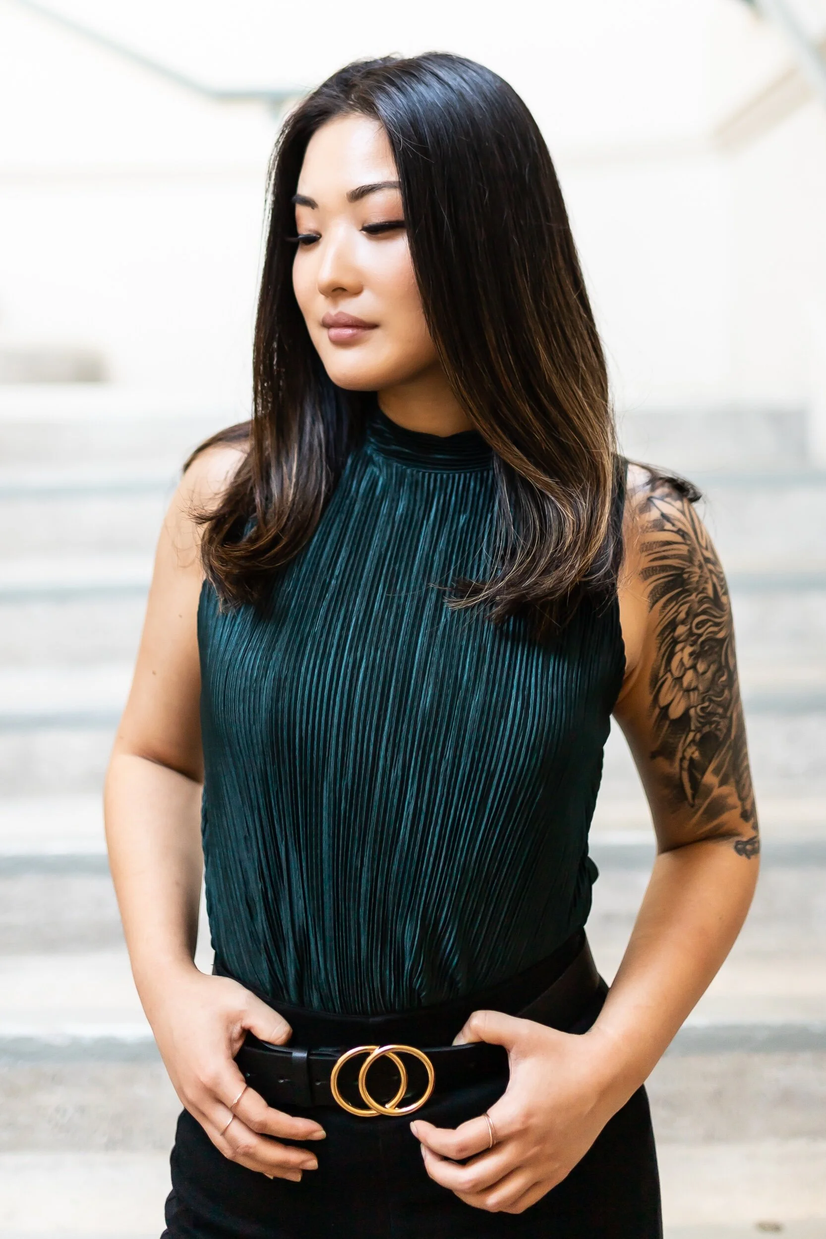 A woman with long dark hair and tattoos on her right arm, wearing a sleeveless dark green top and black pants with a double gold belt buckle, standing outdoors near steps.