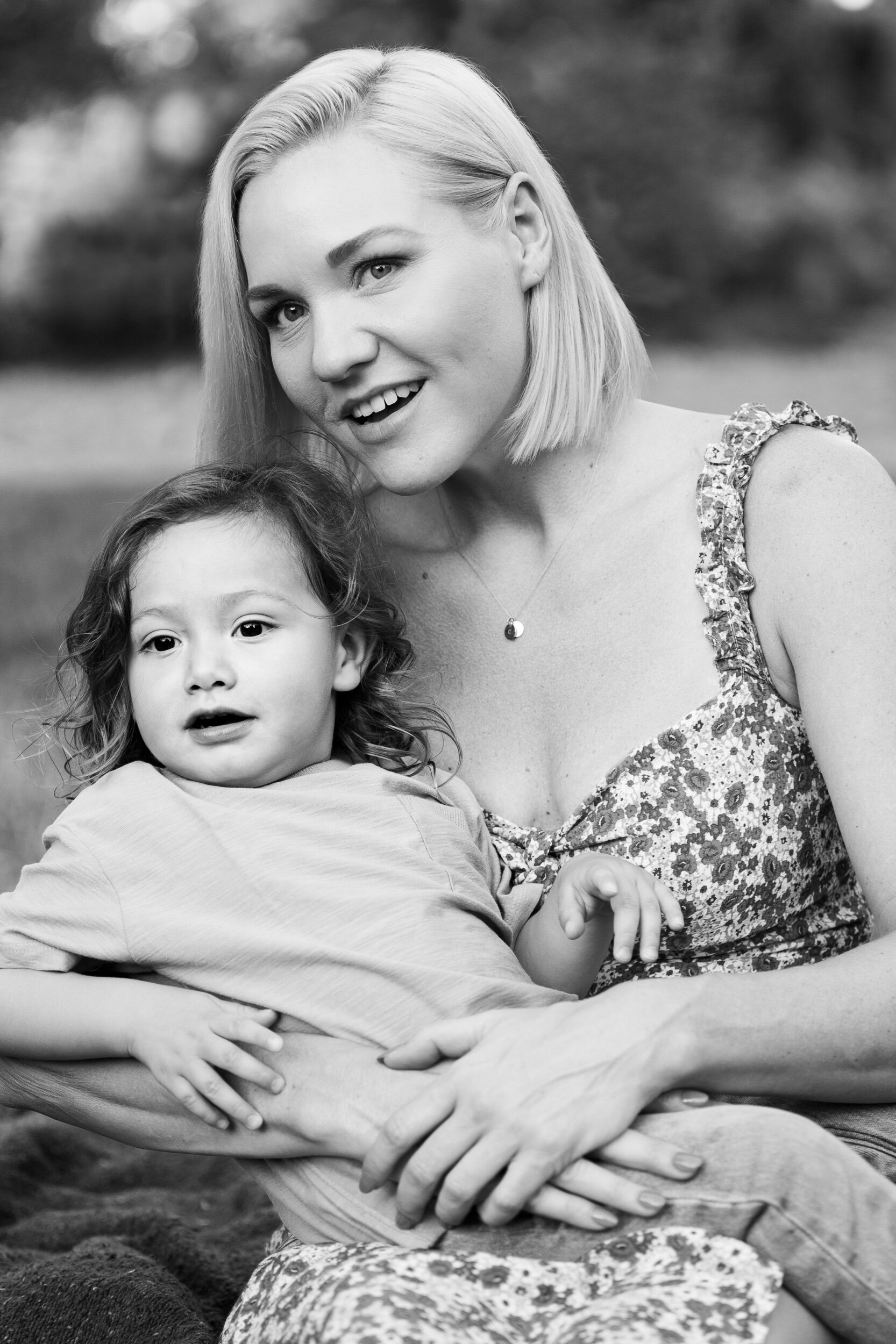 A black and white photo of a smiling woman with blonde hair and a young girl with curly hair, sitting outdoors with blurred natural background.