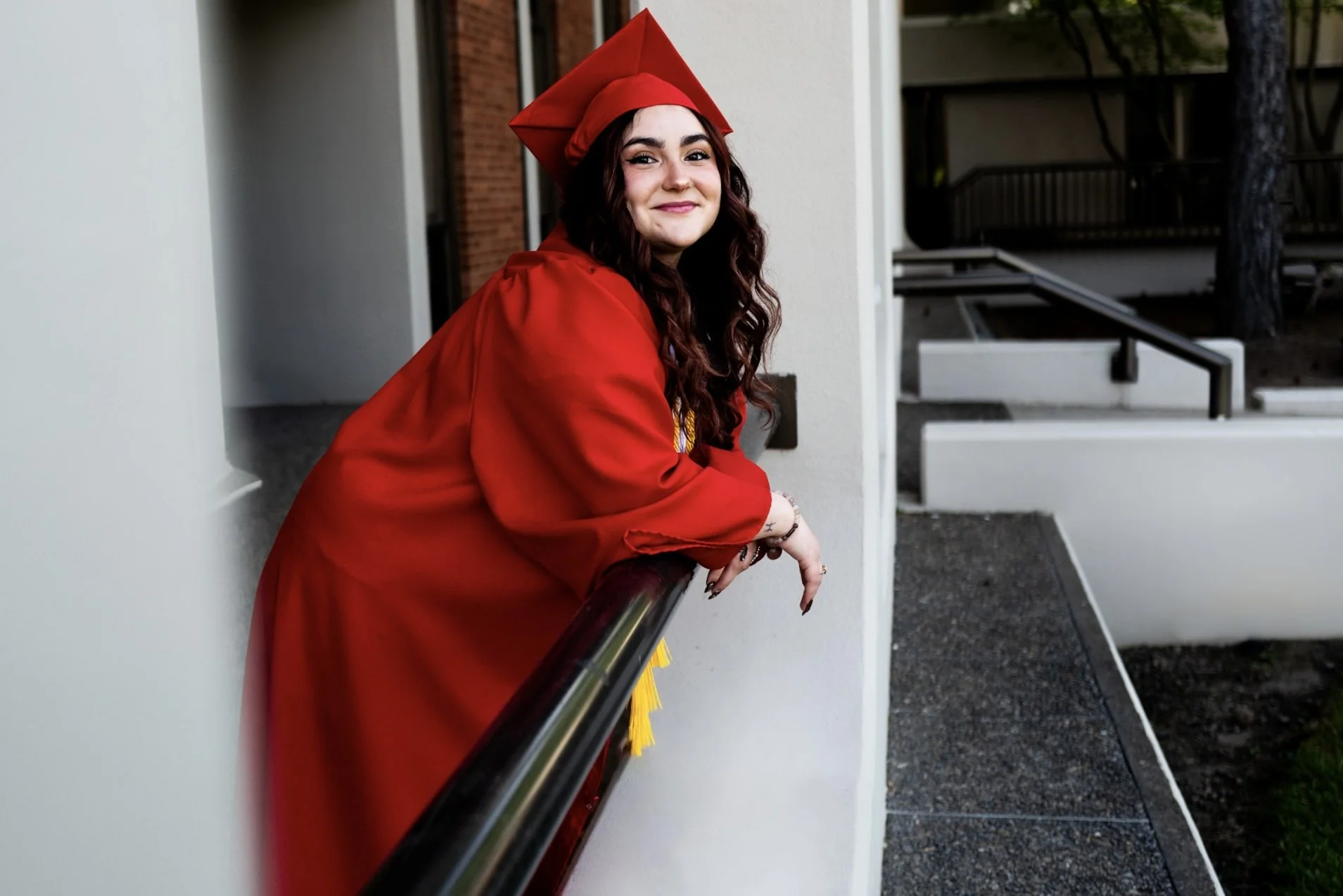 A young woman with long curly hair wearing a red graduation cap and gown, smiling while leaning on a black railing outside a building.