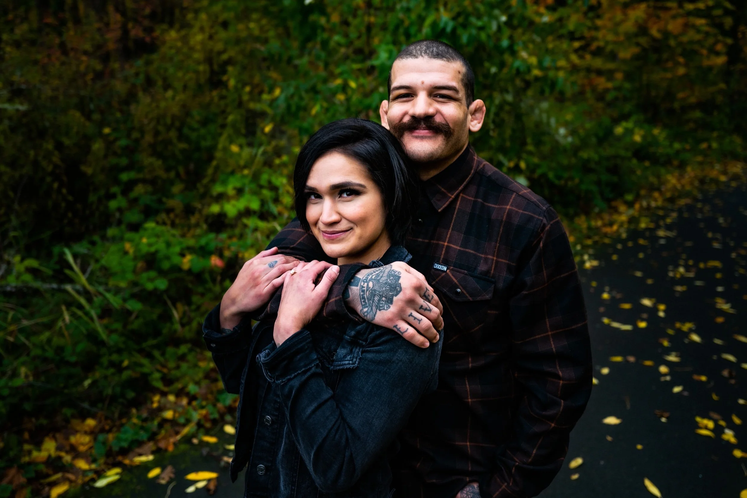 A man and woman are standing outside in a natural setting with greenery and leaves on the ground. The man has short dark hair, a mustache, and tattoos on his hands, and the woman has short black hair and tattoos on her neck. They are embracing, with 