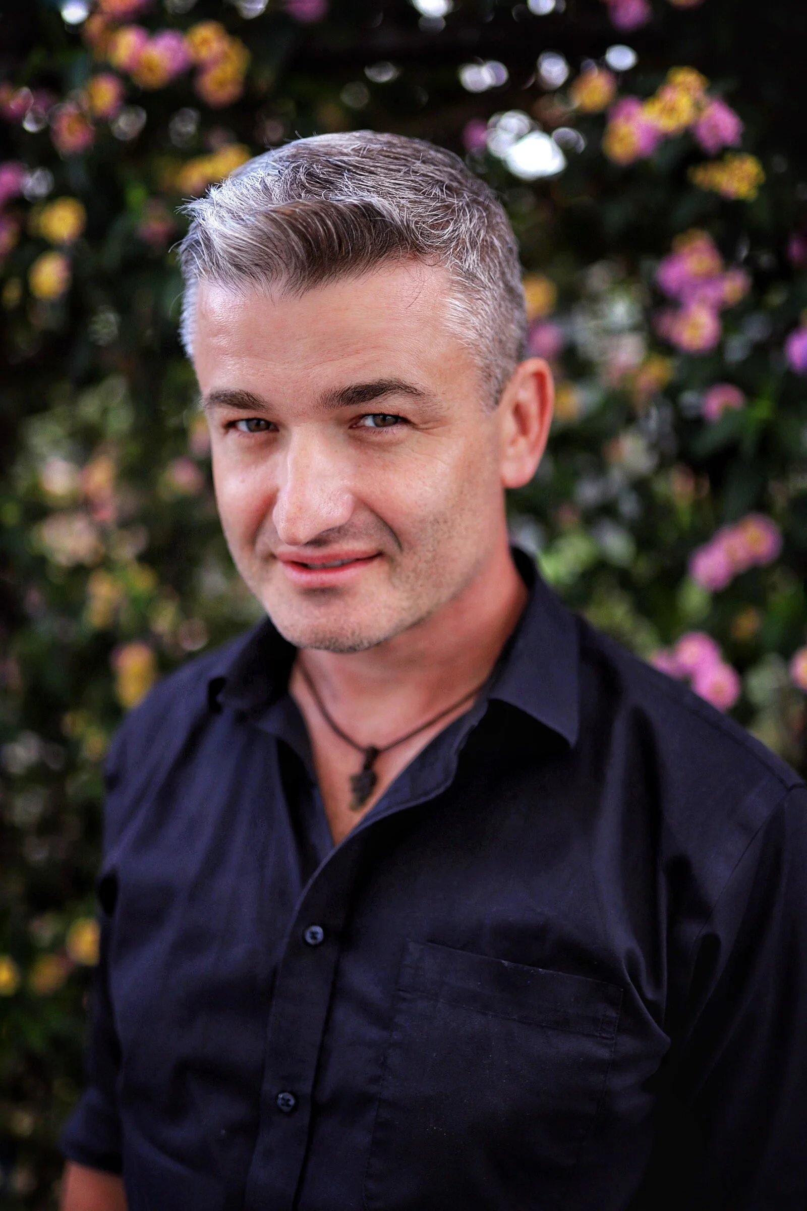 A man with short gray hair and wearing a black shirt, standing outdoors with a background of blurred pink, purple, and yellow flowers.