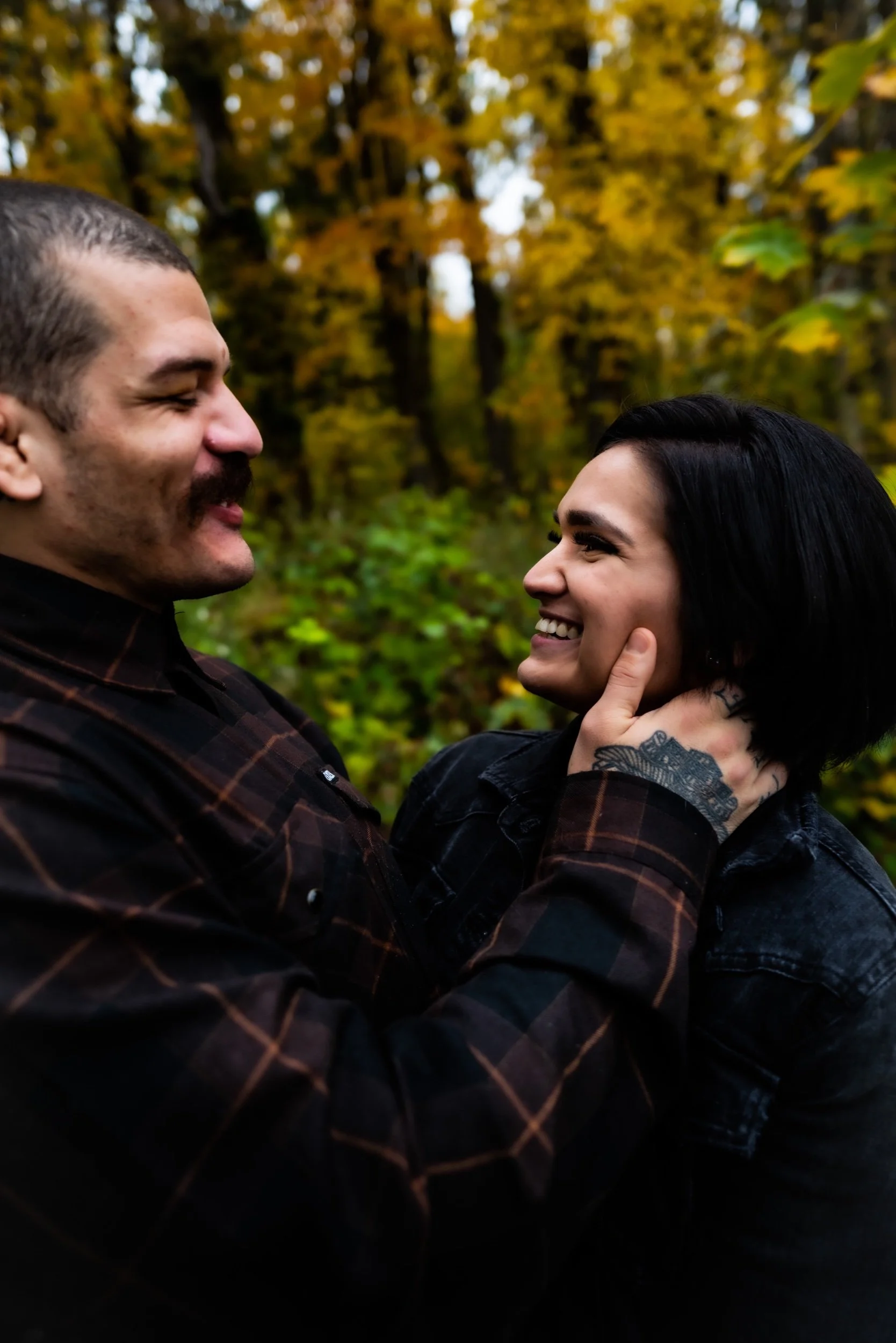 A couple smiling and looking at each other in an outdoor forest setting with autumn foliage.