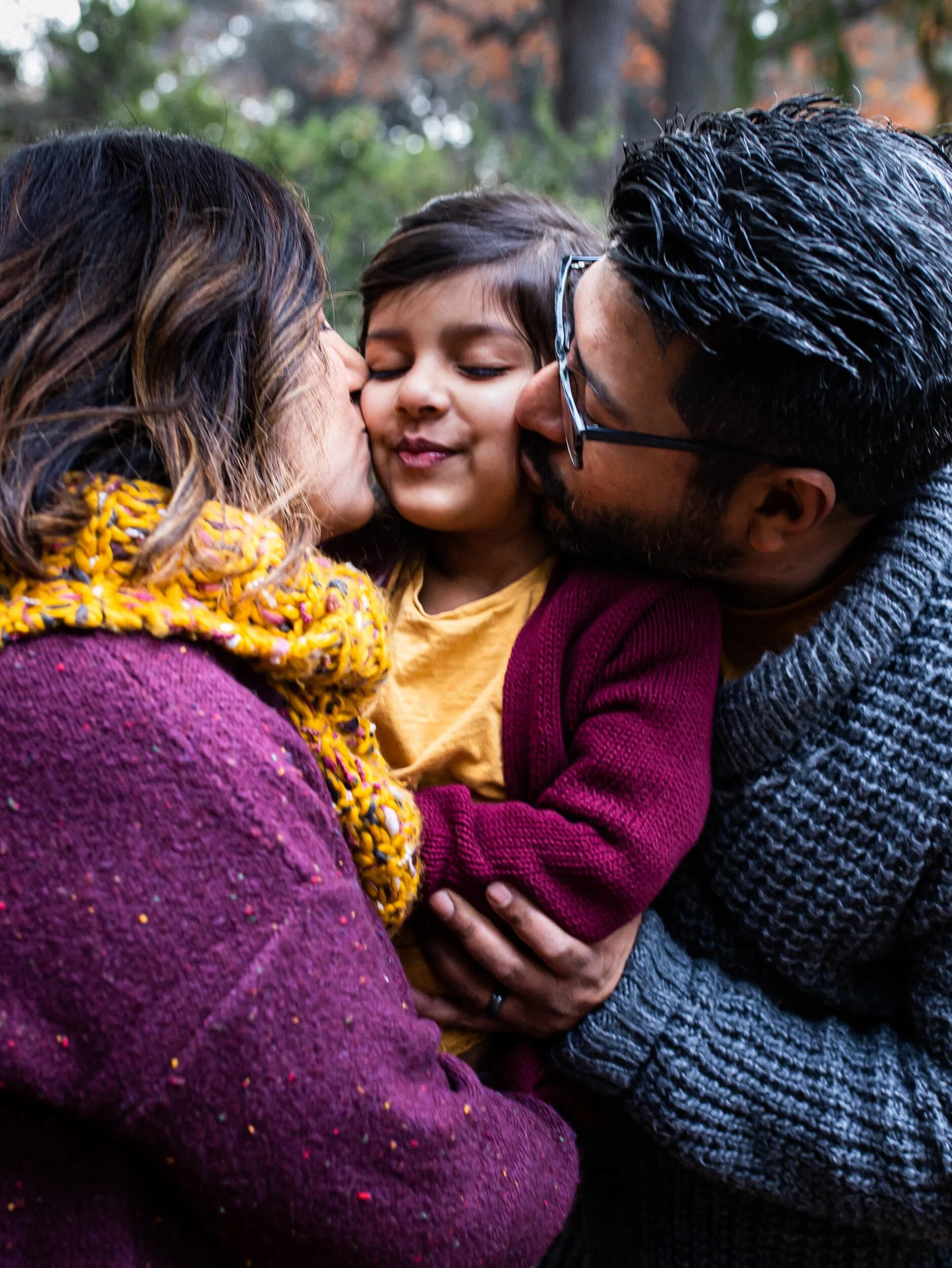 A close-up of a family moment with a woman and man kissing a child's cheeks in an outdoor setting with trees in the background.